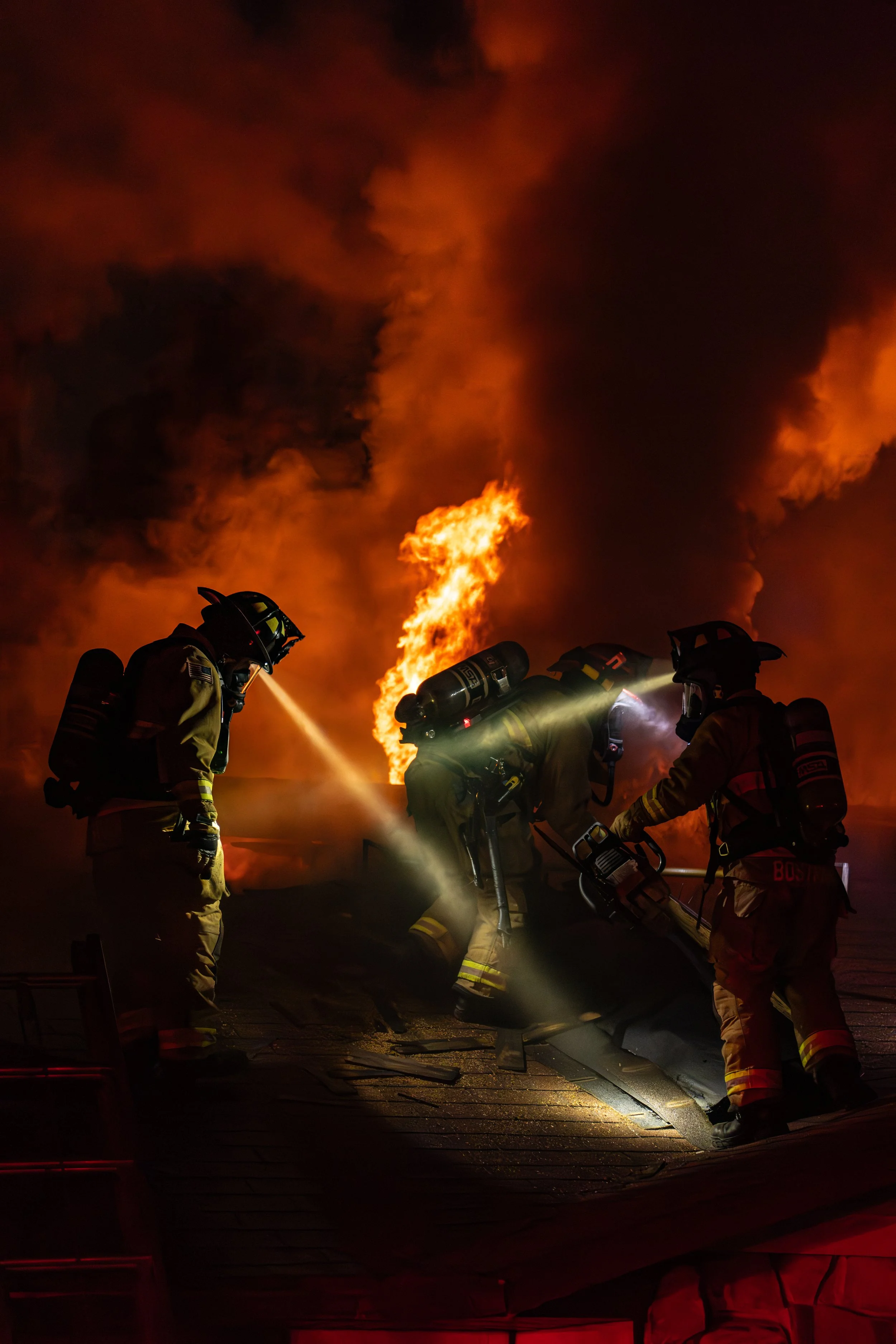 Firefighters in protective gear working to extinguish a large fire, with flames and thick smoke rising in the background at night.