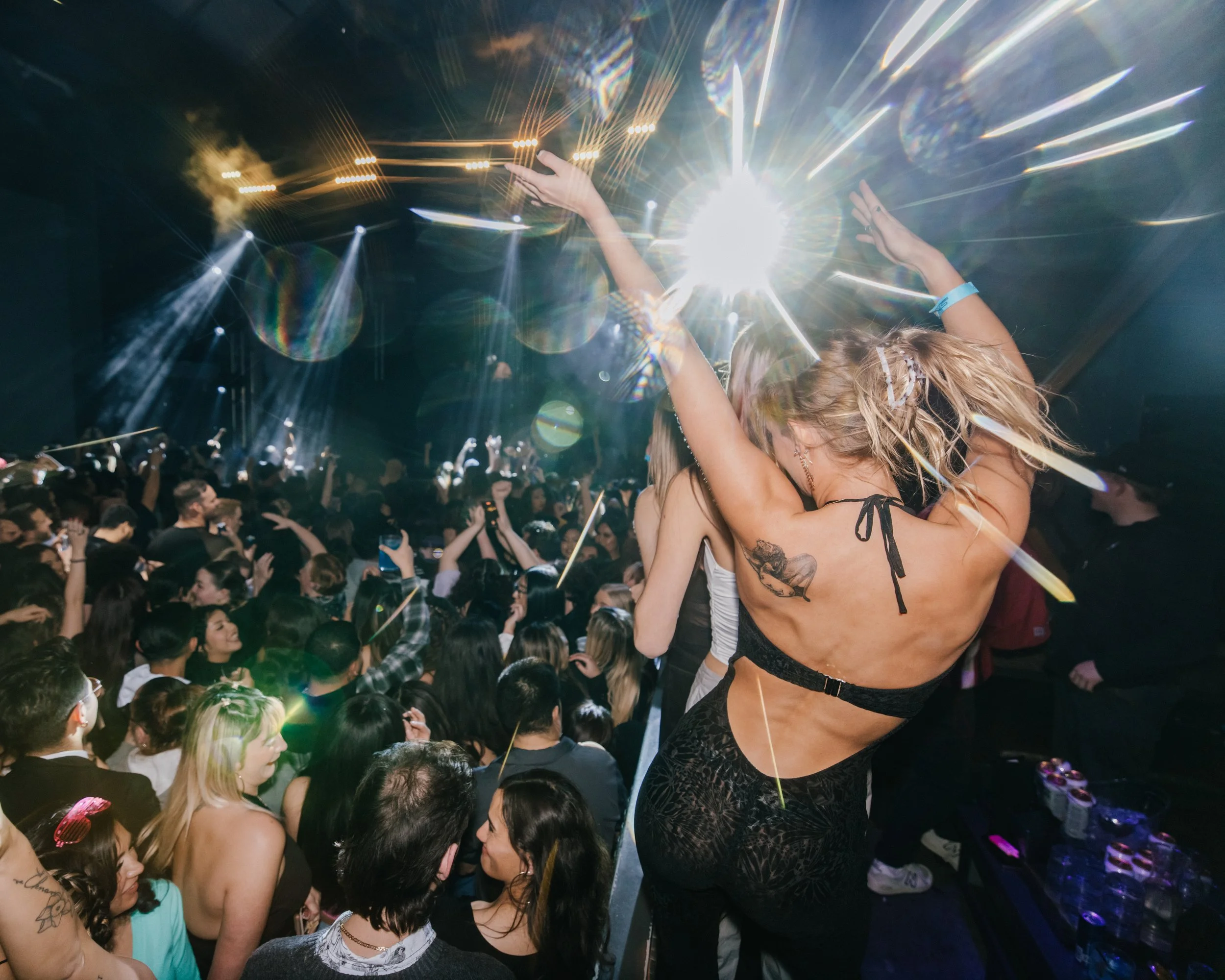 Crowd of people at a concert or nightclub with a woman dancing in the foreground, illuminated by bright stage lights and lens flare.