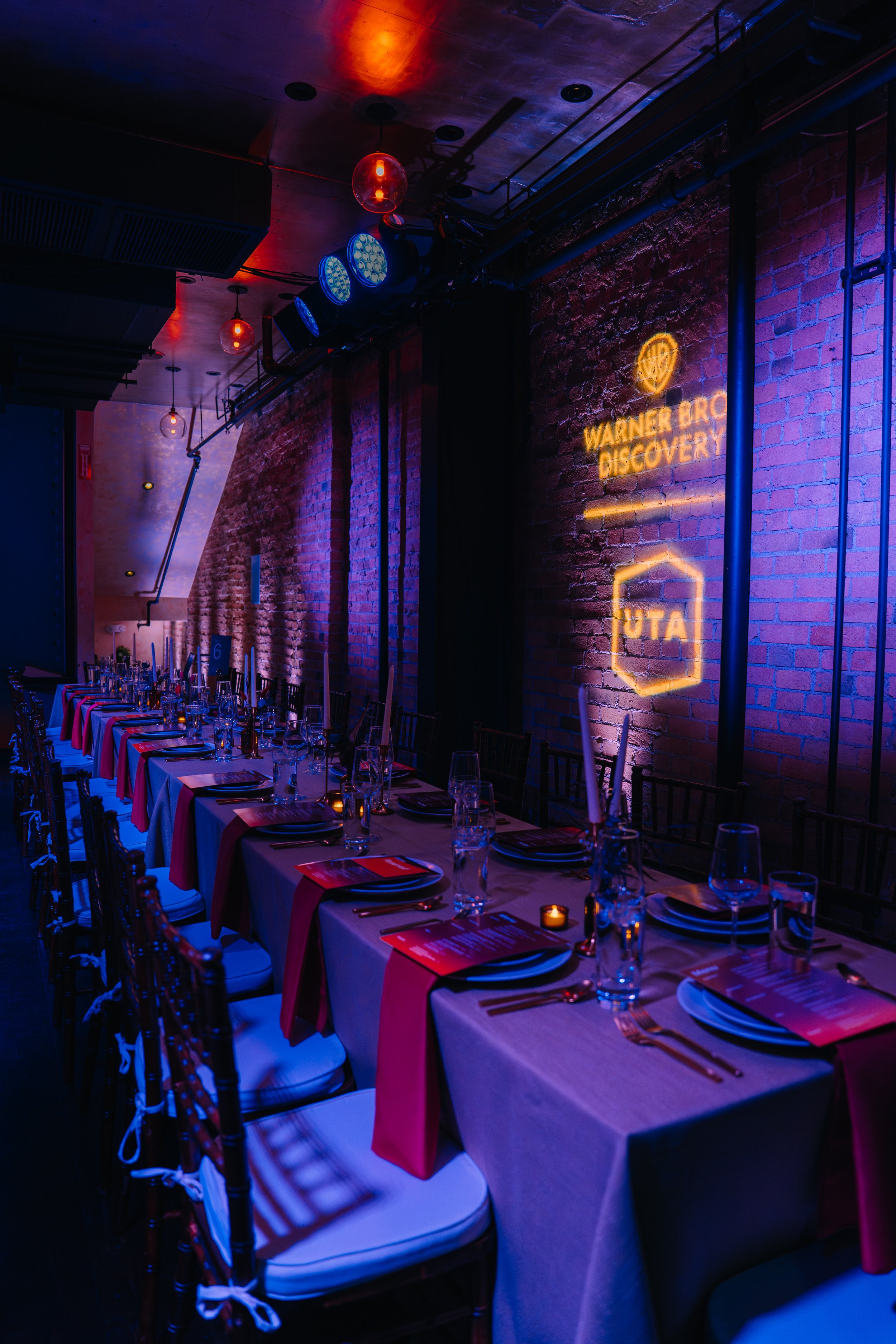 Empty dining table set with glasses, napkins, and utensils in a dimly lit restaurant with red and blue lighting, exposed brick walls, and a neon sign that reads 'Warnet Bro Discovery'.