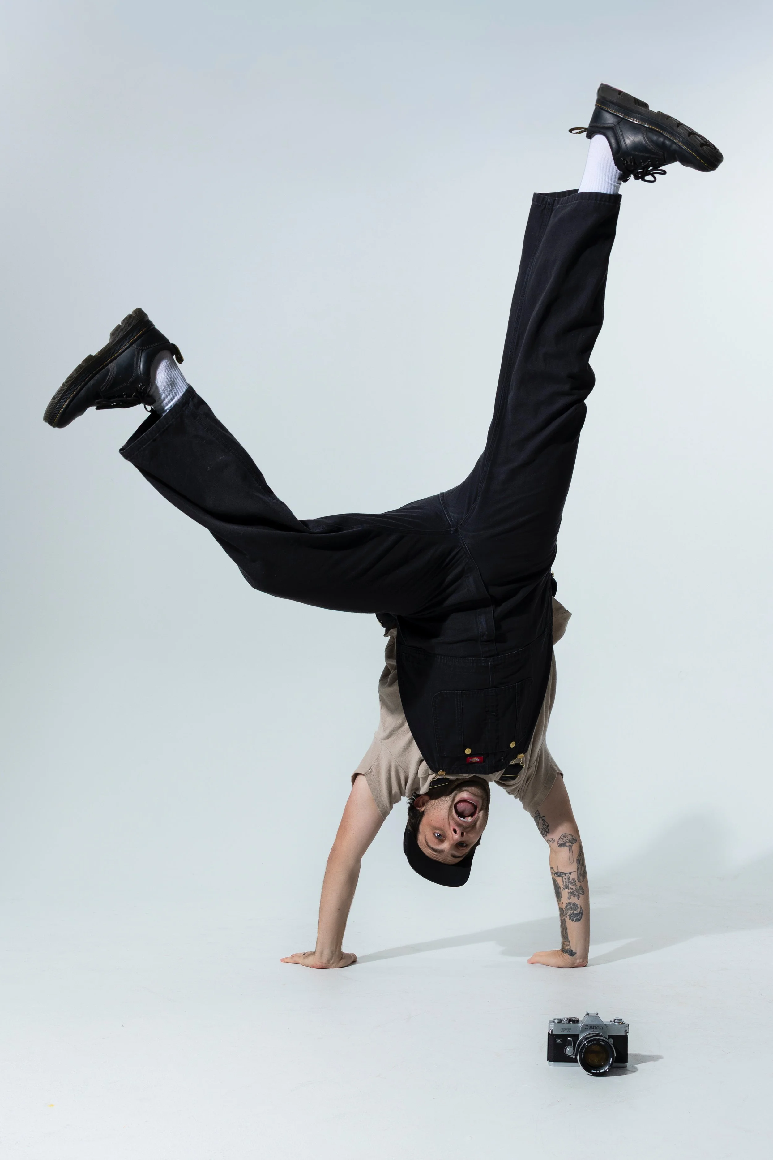 A man wearing a beige shirt, black vest, black cap, and black shoes is performing a handstand on the floor, with his legs in the air. He has tattoos on his right arm and a wide open mouth. There is a camera placed on the floor in front of him, and the background is plain and light-colored.