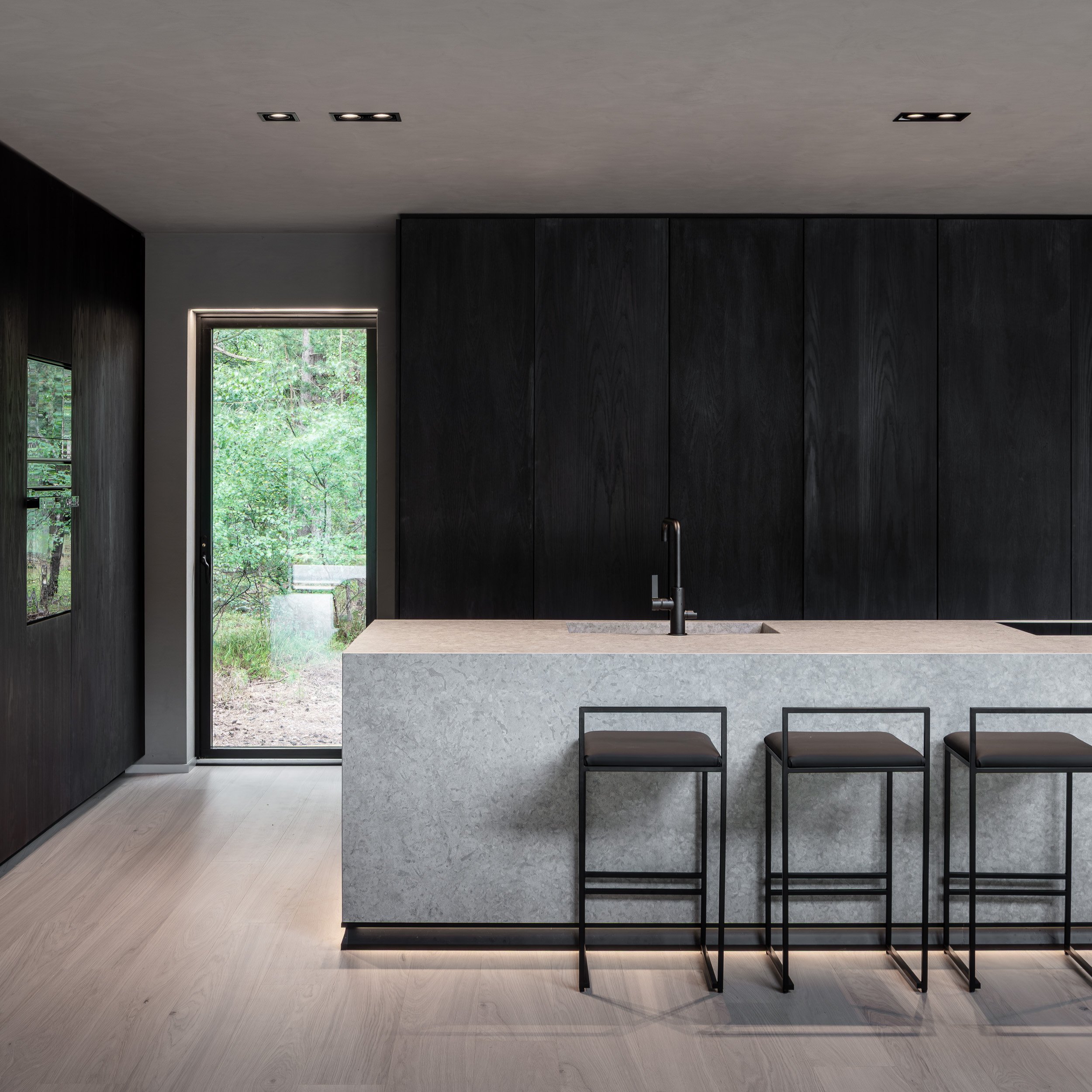 Professionell inredningsfotografering av minimalistiskt stramt kök med svart träinredning, Bricmate-bänkskiva och skogsutsikt genom glasdörr.