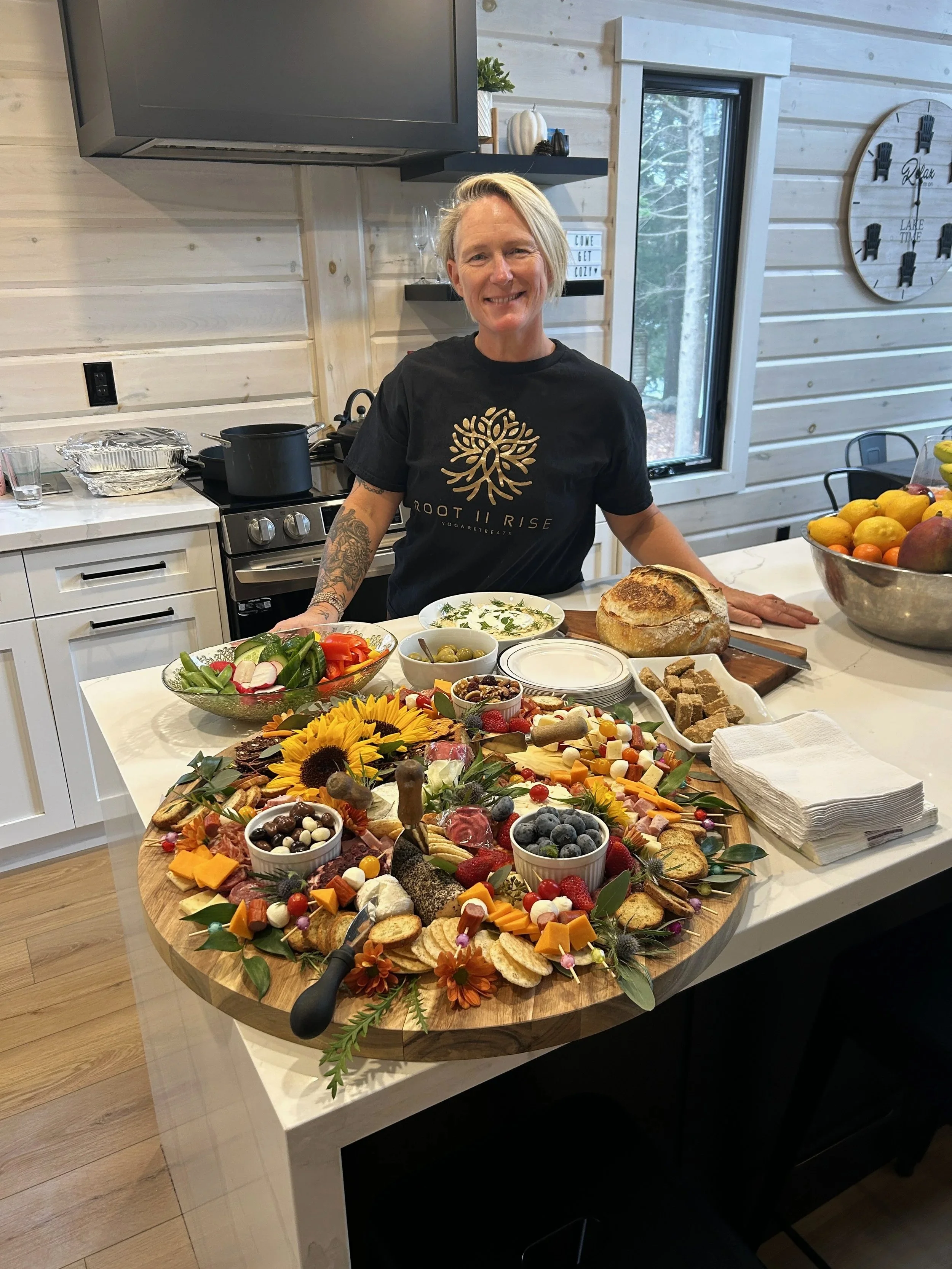 A woman with short blonde hair and tattoos on her arm standing behind a decorated Thanksgiving or fall feast table, smiling. The table holds bread, cheese, fruits, vegetables, and various appetizers. The kitchen features light wood walls, a window, a large bowl of fruit, and a black stove with pots.
