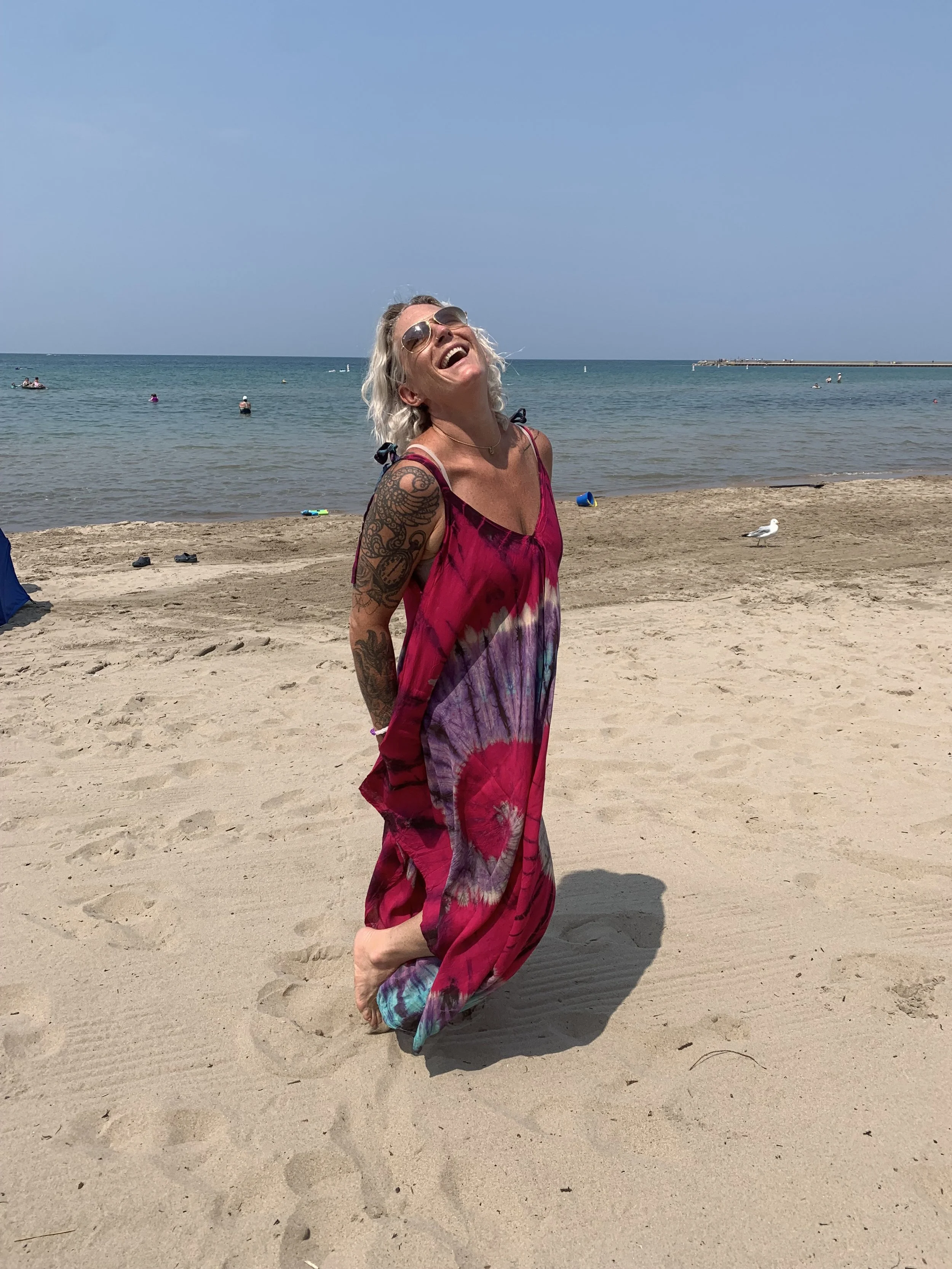 A woman with long curly blonde hair, sunglasses, and a large colorful tattoo on her arm stands barefoot on the sandy beach, laughing and enjoying herself in a flowy pink and purple dress with a background of the ocean and clear blue sky.