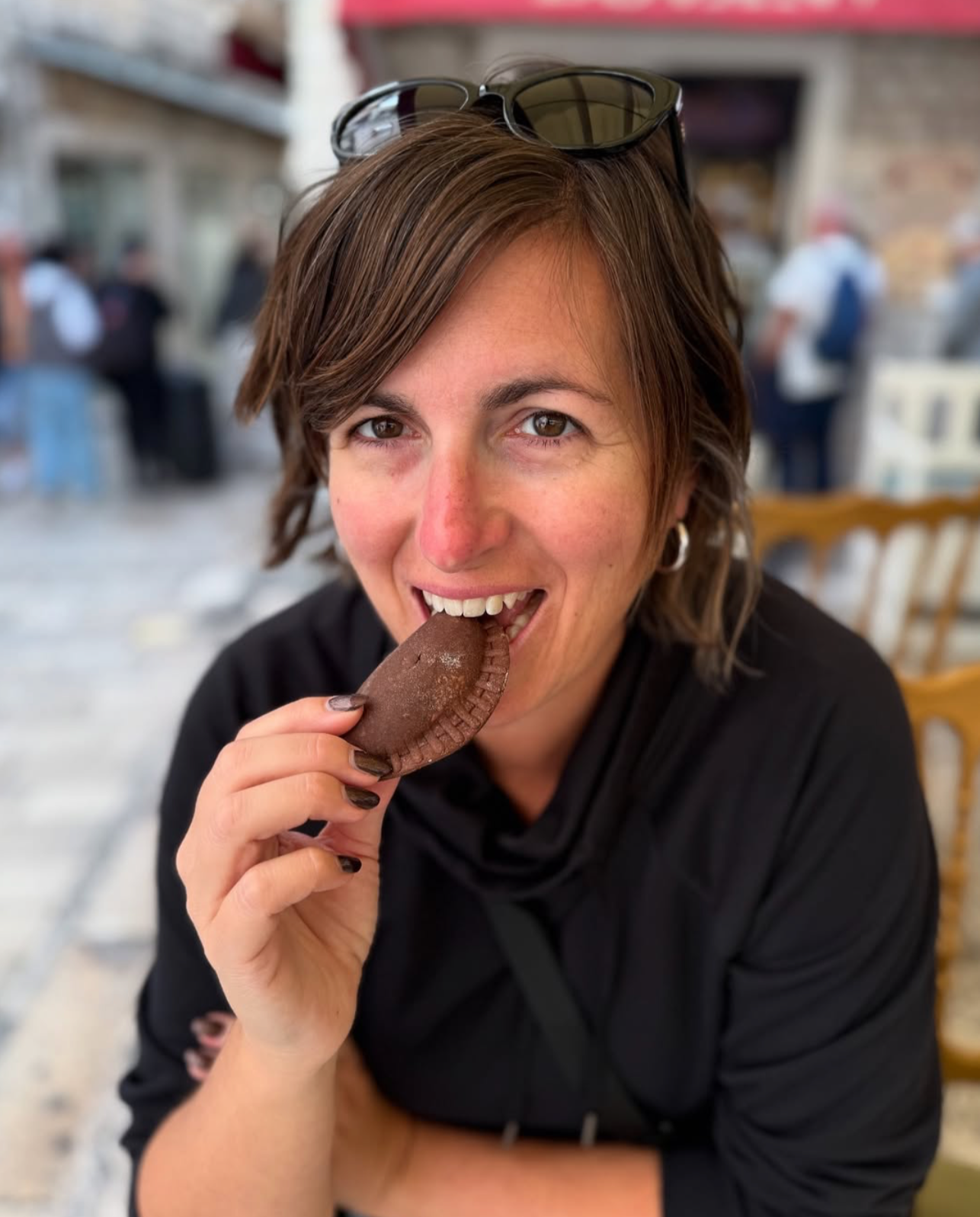 A woman with short brown hair and sunglasses on her head is smiling and holding a brown empanada near her mouth. She is sitting outdoors with a blurred background of people and buildings.