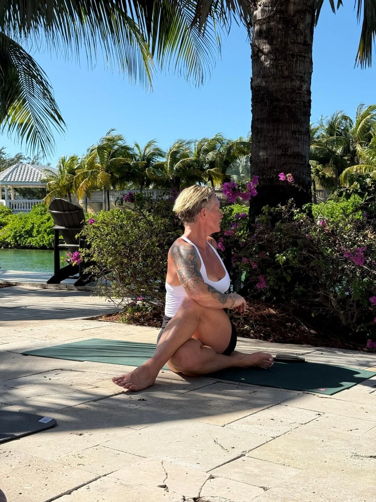 A woman with tattoos practicing yoga outdoors on a mat near a palm tree and pink flowering bushes, with a body of water and a gazebo in the background.