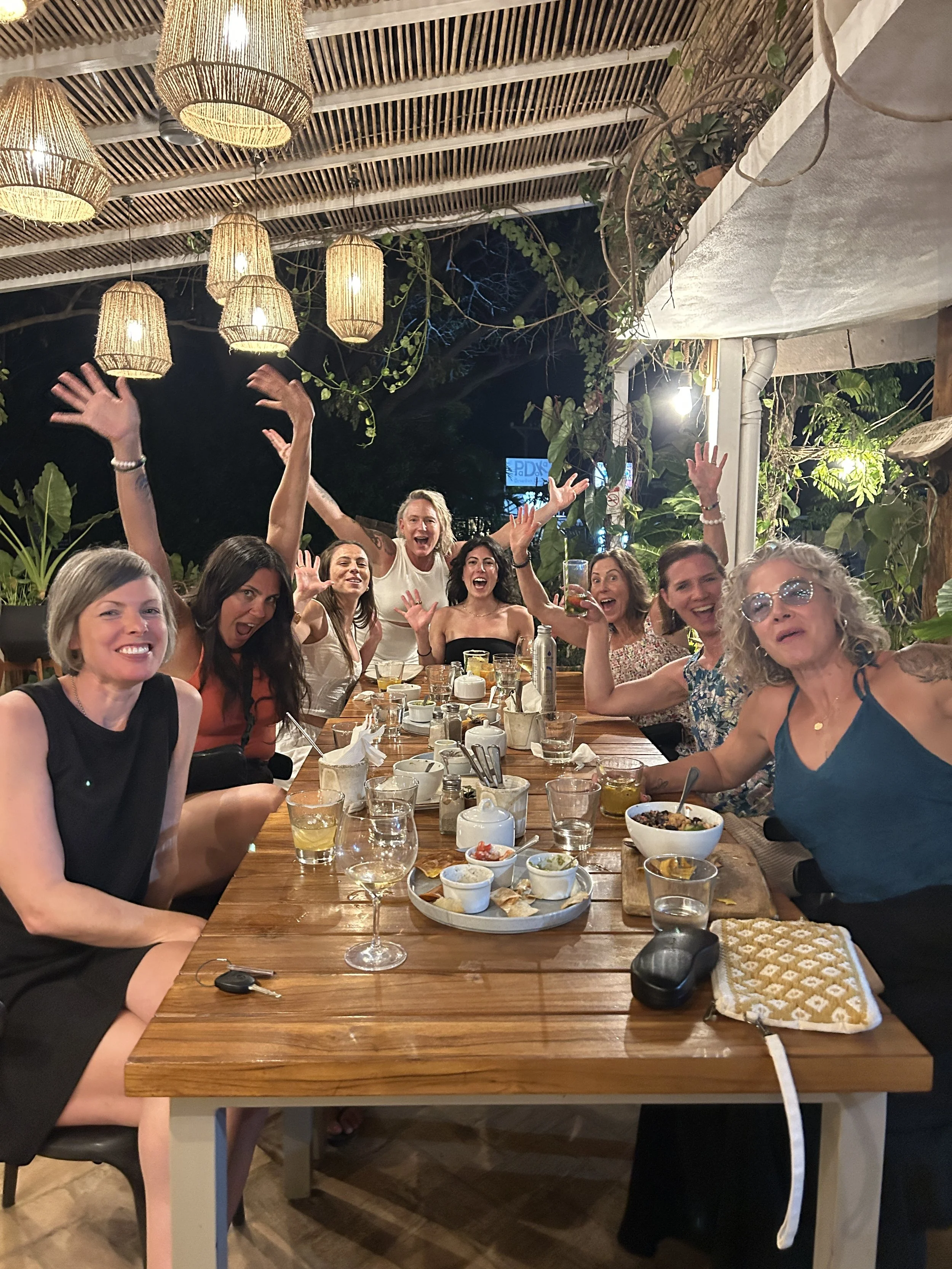 A group of women sitting at a wooden table enjoying a meal and celebrating, with some raising their hands and smiling, in an outdoor patio with hanging lights and plants.