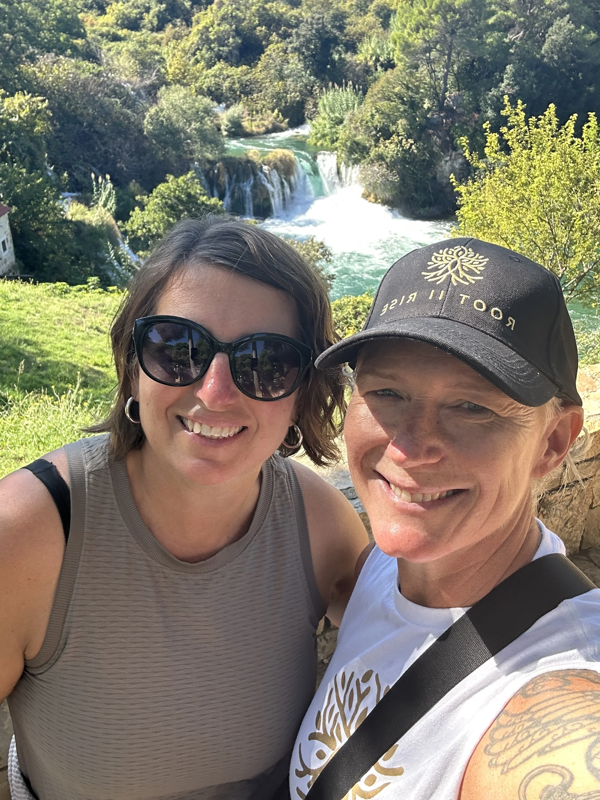 A selfie of a smiling woman wearing sunglasses and a sleeveless top, and a man wearing a baseball cap and a white t-shirt, with a lush green landscape, waterfall, and trees in the background.