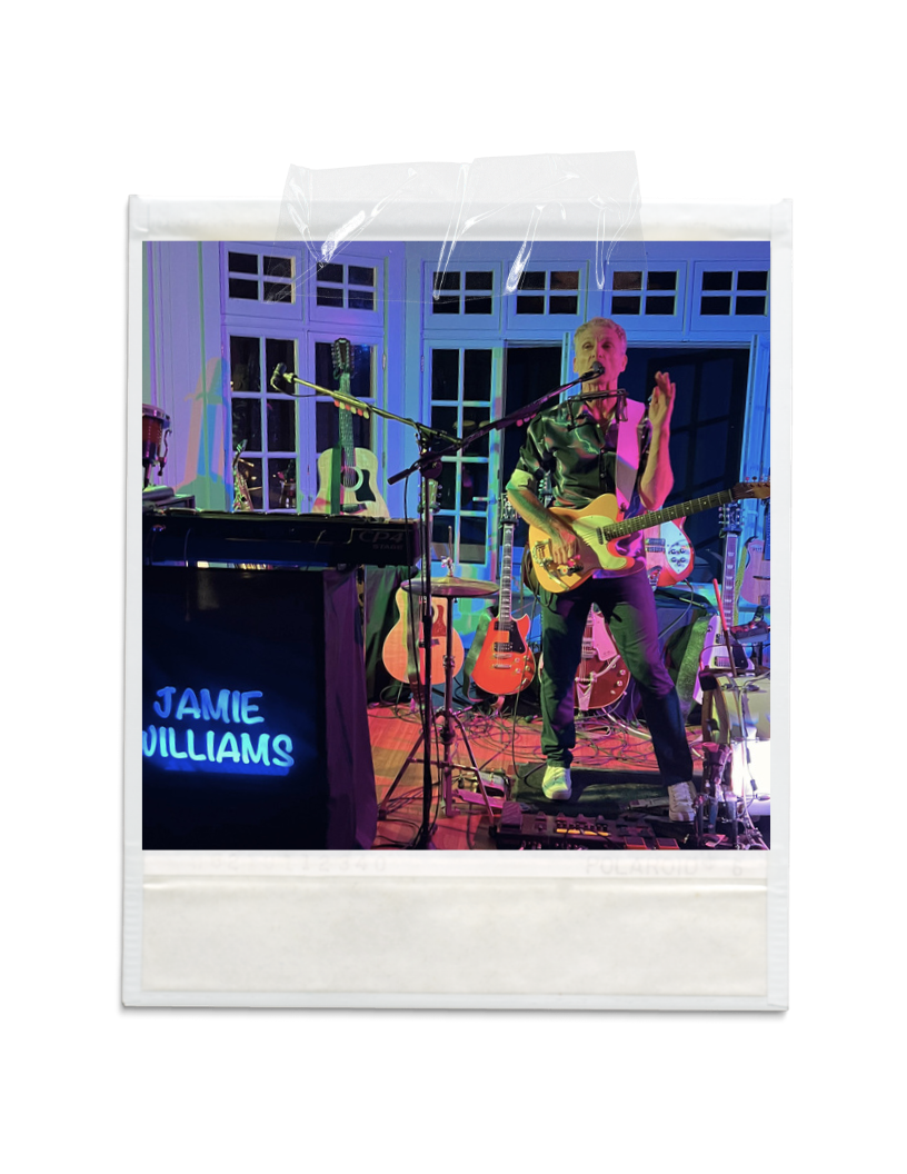 A musician performing on stage with multiple guitars in the background, illuminated by colorful lighting, and a neon sign that reads 'JAMIE WILLIAMS'.
