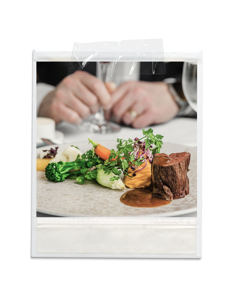 Close-up of a gourmet dish with a beef filet, vegetables, and sauce on a plate, with a person in the background taking a photo of it.