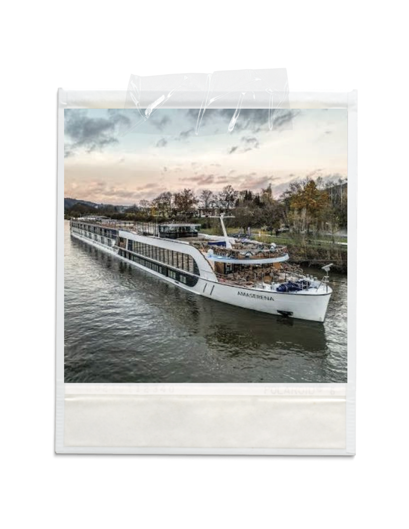 A large white boat named 'Amserena' is cruising on a river with trees and buildings in the background during cloudy weather.