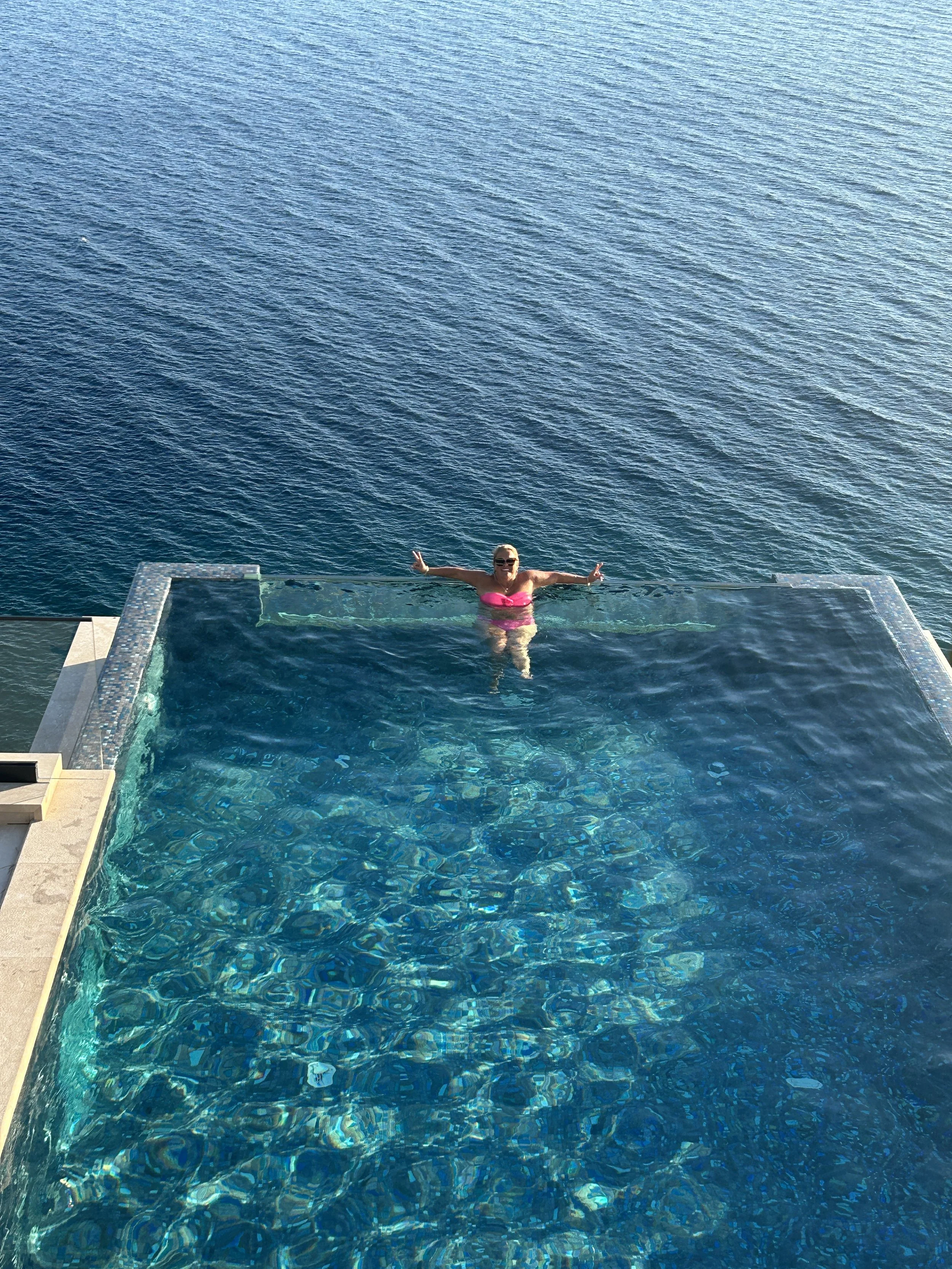 Woman in pink swimsuit and sunglasses relaxing in an infinity pool overlooking the ocean.