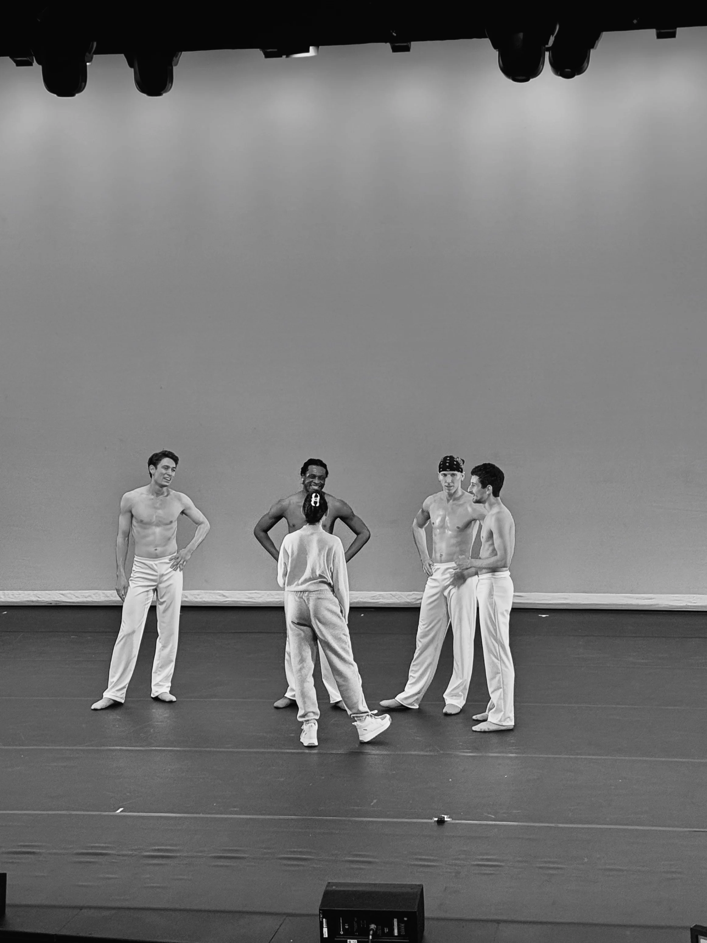 Five performers on stage, four shirtless men and one woman in casual wear, engaged in a conversation, with a plain background and stage lighting overhead.