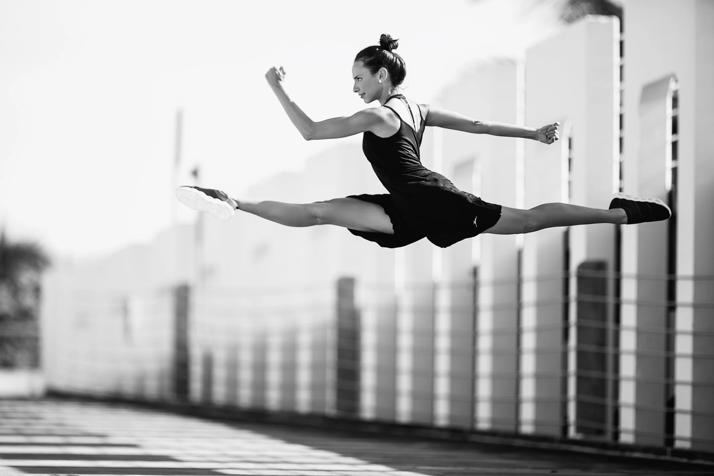 A woman in athletic clothing performs a high jump leap outdoors in front of a modern building, captured in black and white.