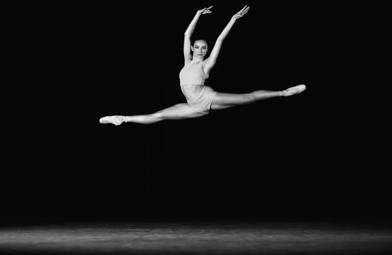 A female ballet dancer performing a grand jeté leap on stage in front of a dark background.