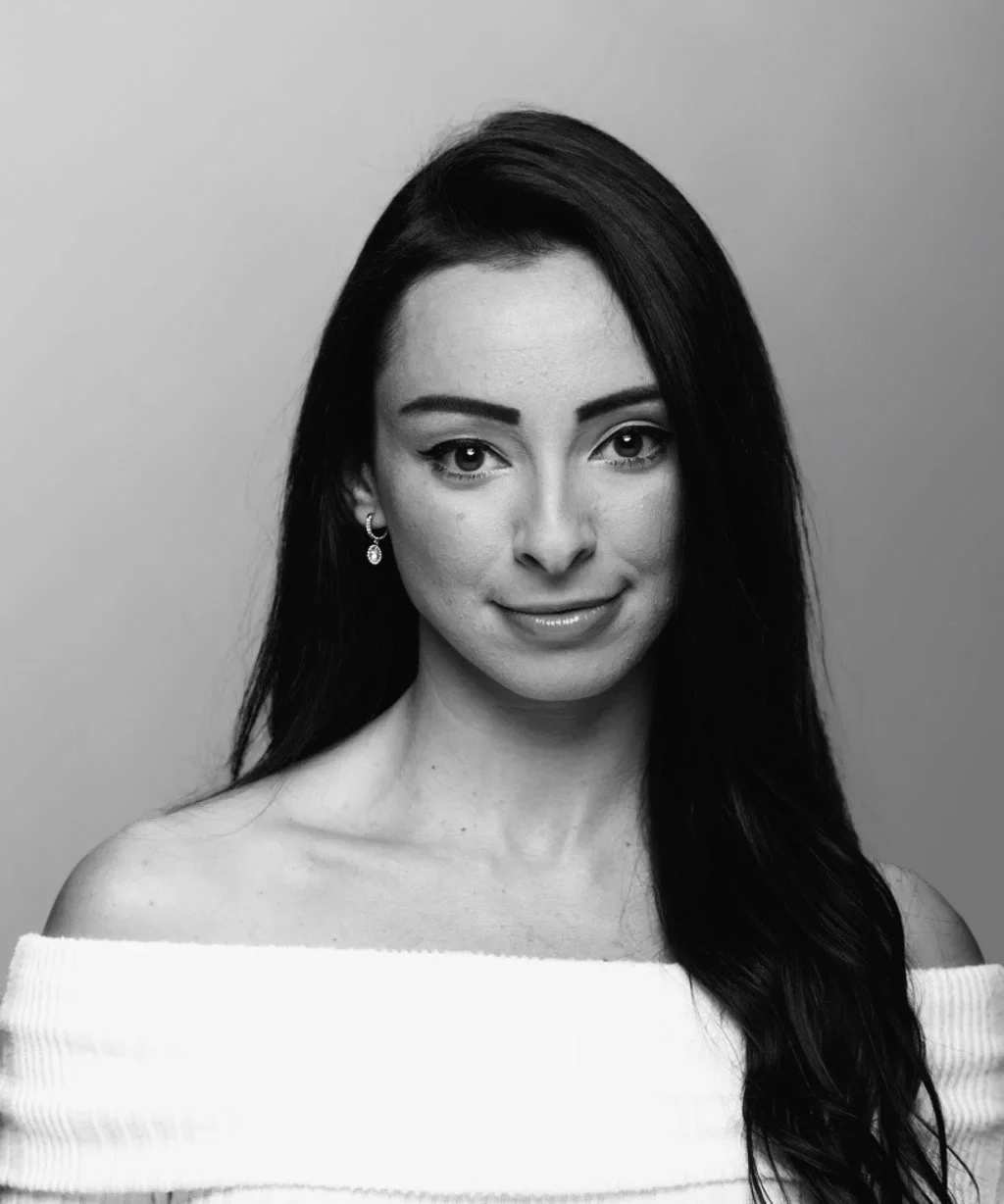 Black and white portrait of a woman with long dark hair, wearing earrings and an off-the-shoulder top, looking at the camera with a slight smile.