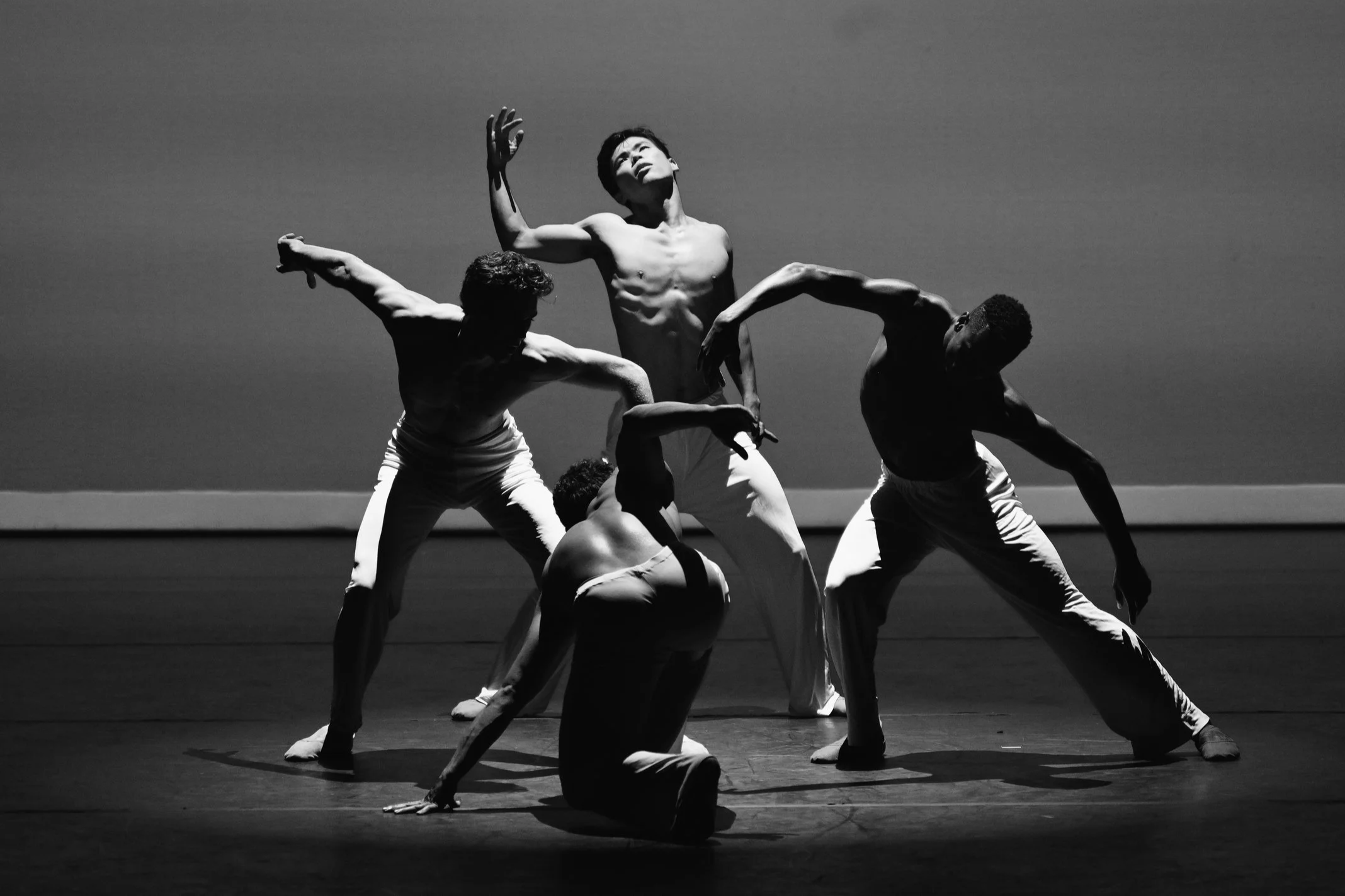 Black and white photo of five male dancers performing on stage, with dramatic lighting highlighting their muscular bodies and expressive poses.