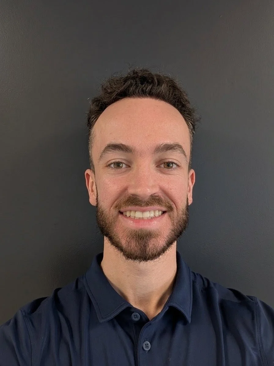 A young man with short curly dark hair, a beard, and a big smile, wearing a dark blue collared shirt, standing against a plain dark wall.
