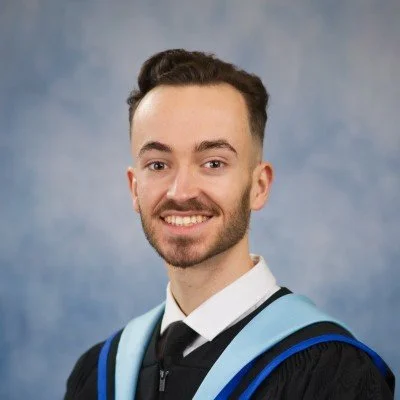 Young man in graduation gown and cap smiling, standing against a blue background.