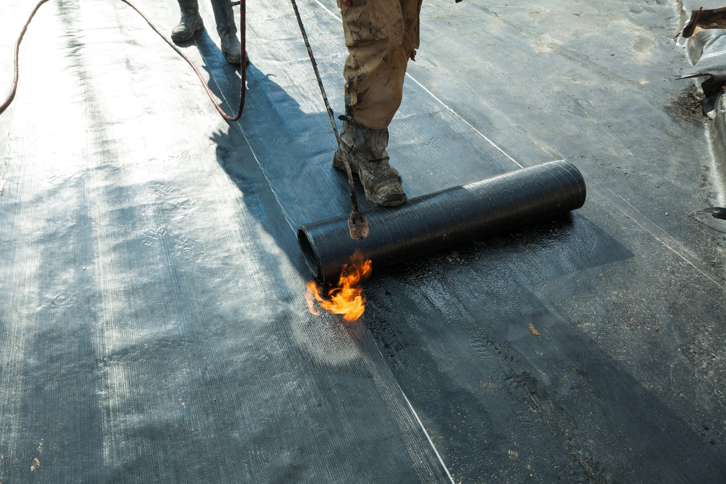 Workers installing a black waterproof membrane on a flat surface, with one worker rolling out the membrane while sparks are coming from a torch underneath the roll.