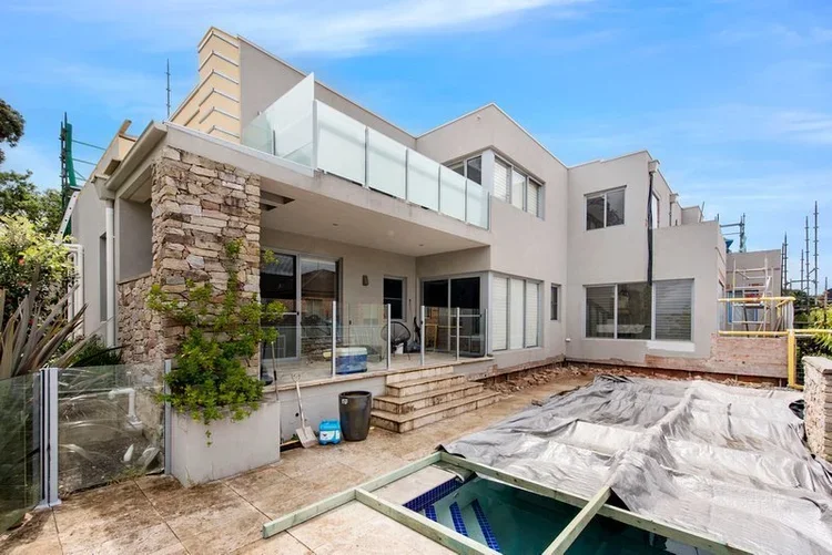 Modern multi-story house under construction with sliding glass doors, a balcony with glass railing, a stone column, and a backyard with a pool under a tarp and construction materials.