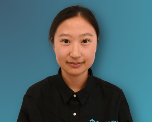 A woman with dark hair pulled back, wearing a black collared shirt with a logo, stands against a solid blue background.