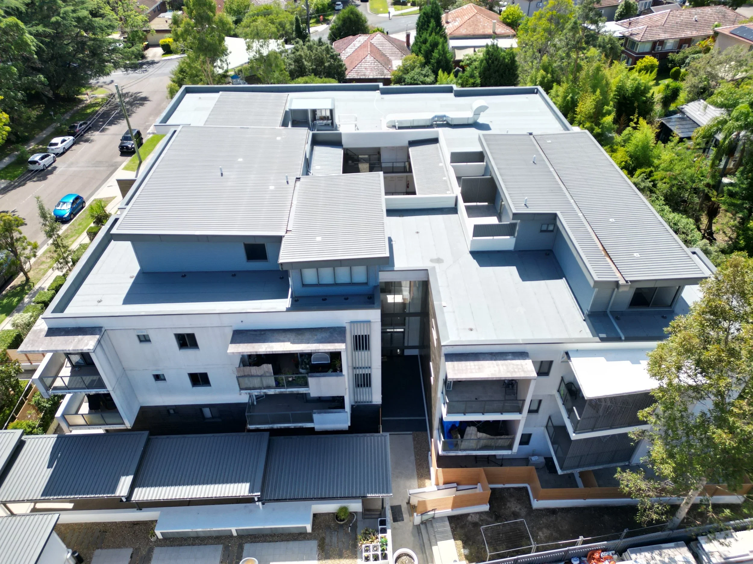 Aerial view of a modern white apartment building with flat roofs and balconies, surrounded by trees, other residential houses, and parked cars on the street.