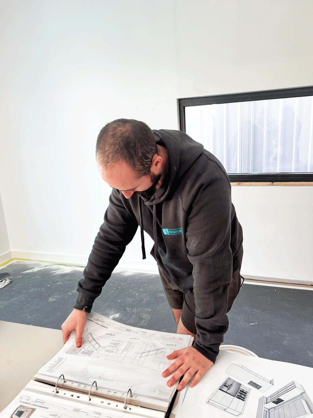 A man looking down at an architectural blueprint on a table, in a room with white walls and a window.