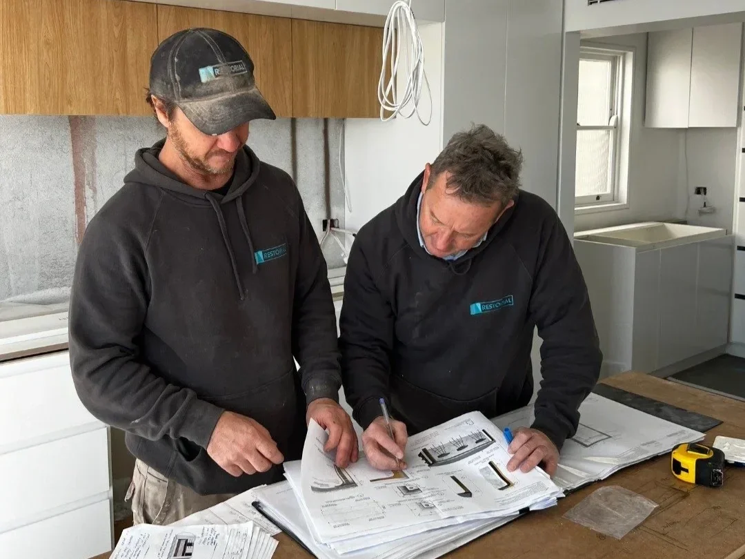 Two men in black hoodies working on construction blueprints in a partially renovated kitchen.