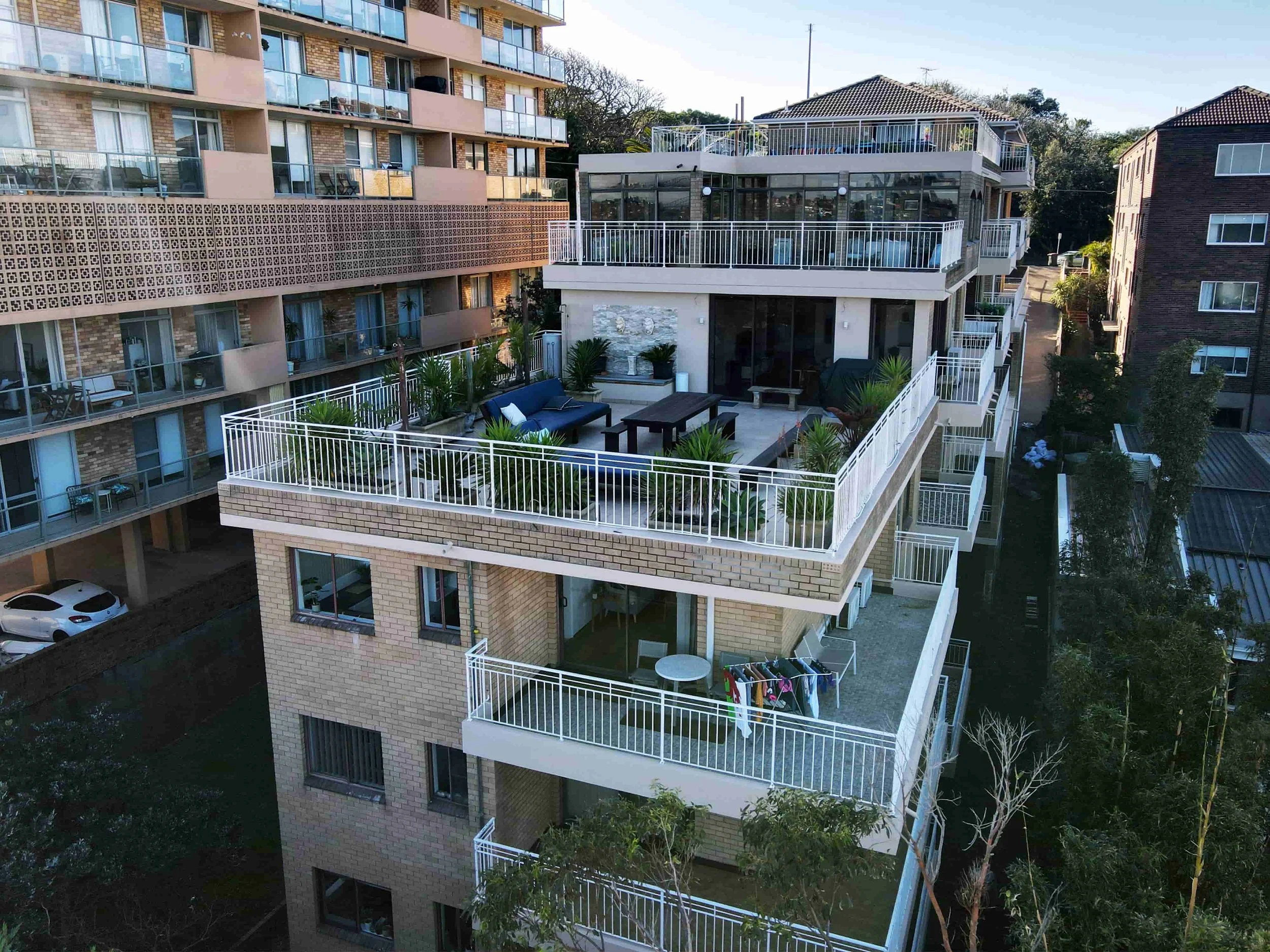 Tall apartment building with multiple balconies, some with outdoor furniture, and a rooftop terrace. The building features a mix of brick and beige exterior with glass railings. Surrounding trees and neighboring buildings are visible.
