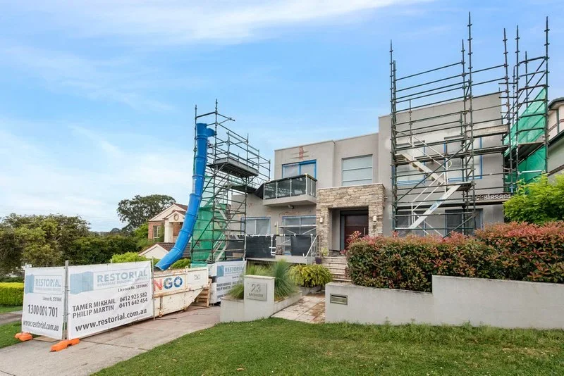 Modern house under construction with scaffolding on the front, a blue construction chute, bushes, and a lawn.