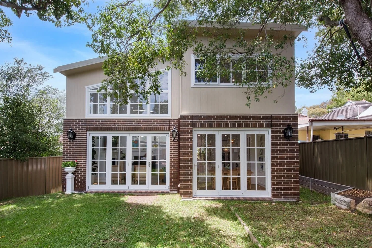 Backyard view of a two-story house with brick and beige siding, large white-framed sliding glass doors on the ground level, white-framed windows on the upper level, surrounded by green grass, trees, and a wooden fence.