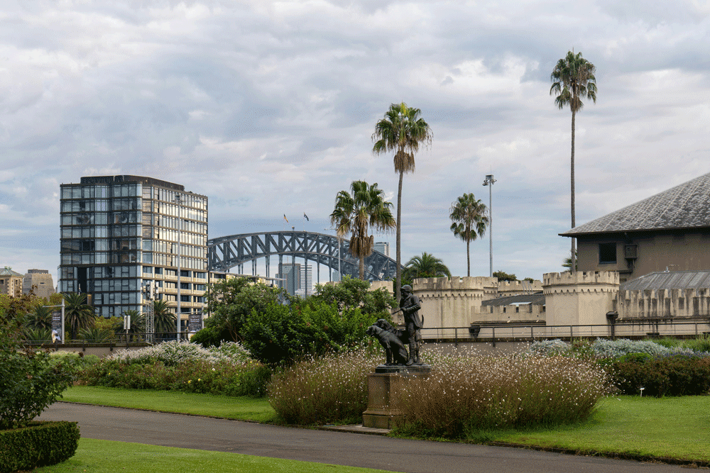 A cityscape with a grassy park area, statues, palm trees, modern buildings, and a large bridge in the background under a cloudy sky.