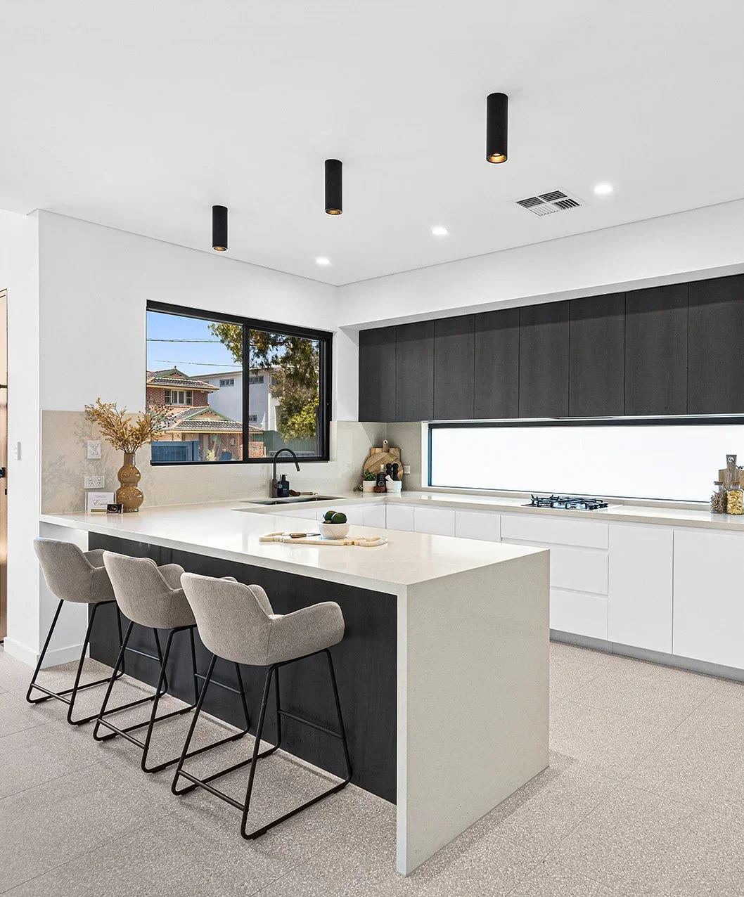 Modern kitchen with white countertops, black and dark gray cabinetry, a large window, and three beige barstools at a kitchen island.