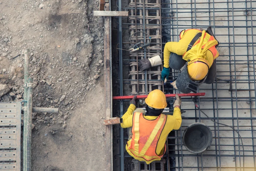 Two construction workers wearing yellow helmets and safety vests working on rebar reinforcement for concrete at a construction site.