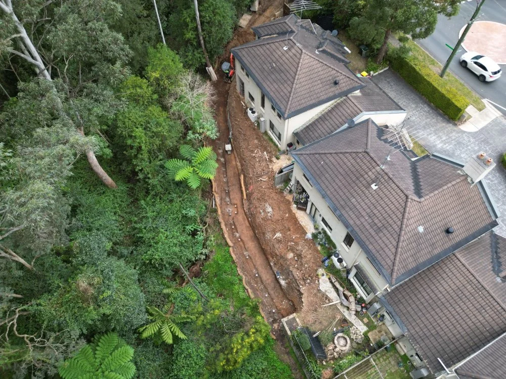 Aerial view of construction work on a house with a trench dug alongside the building, surrounded by trees and a street with parked cars.