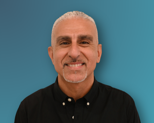 A smiling man with short gray hair and a goatee, wearing a black collared shirt, standing against a blue background.