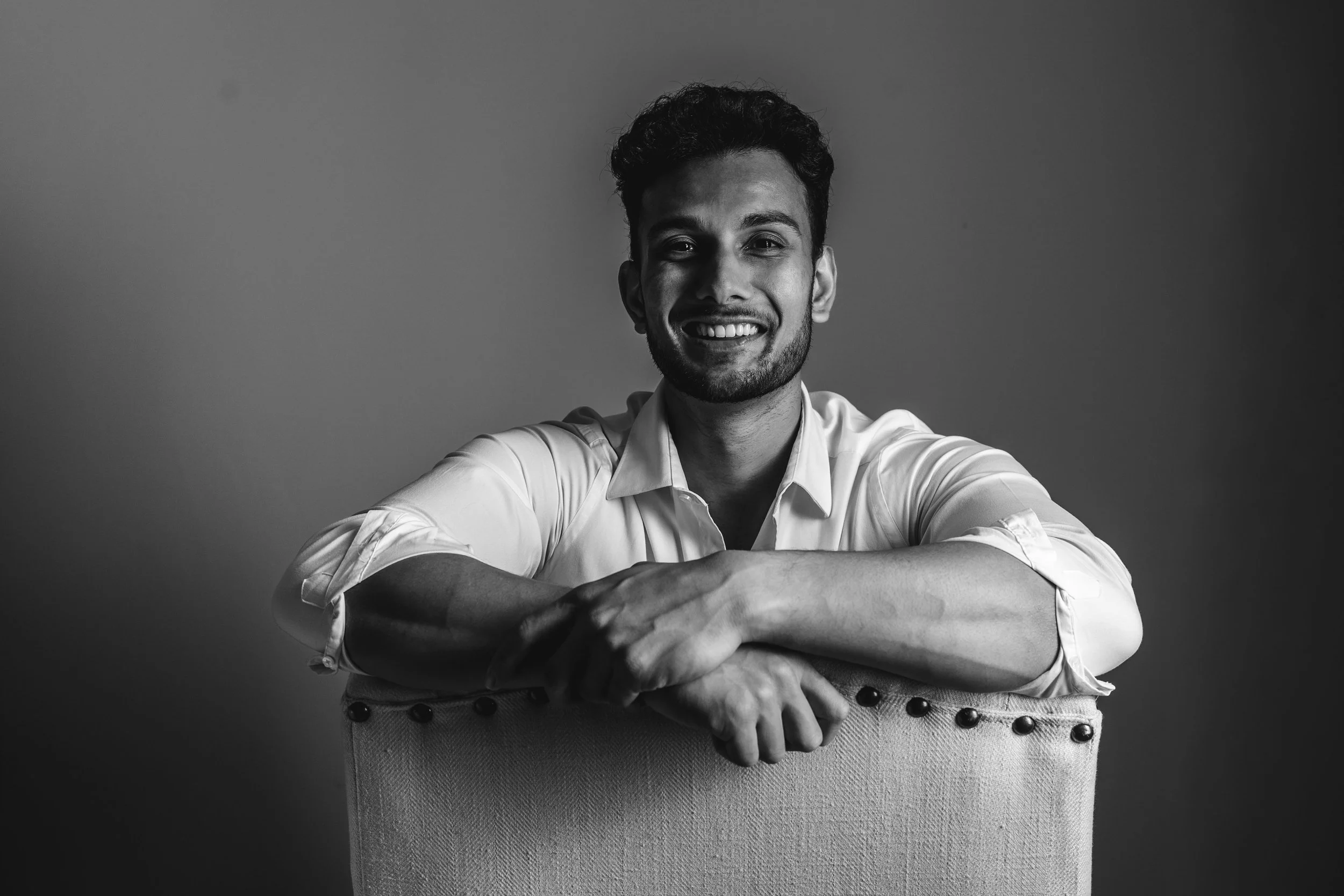 Black and white photo of a smiling man with short hair and a beard, sitting on a chair with arms crossed, against a plain background.
