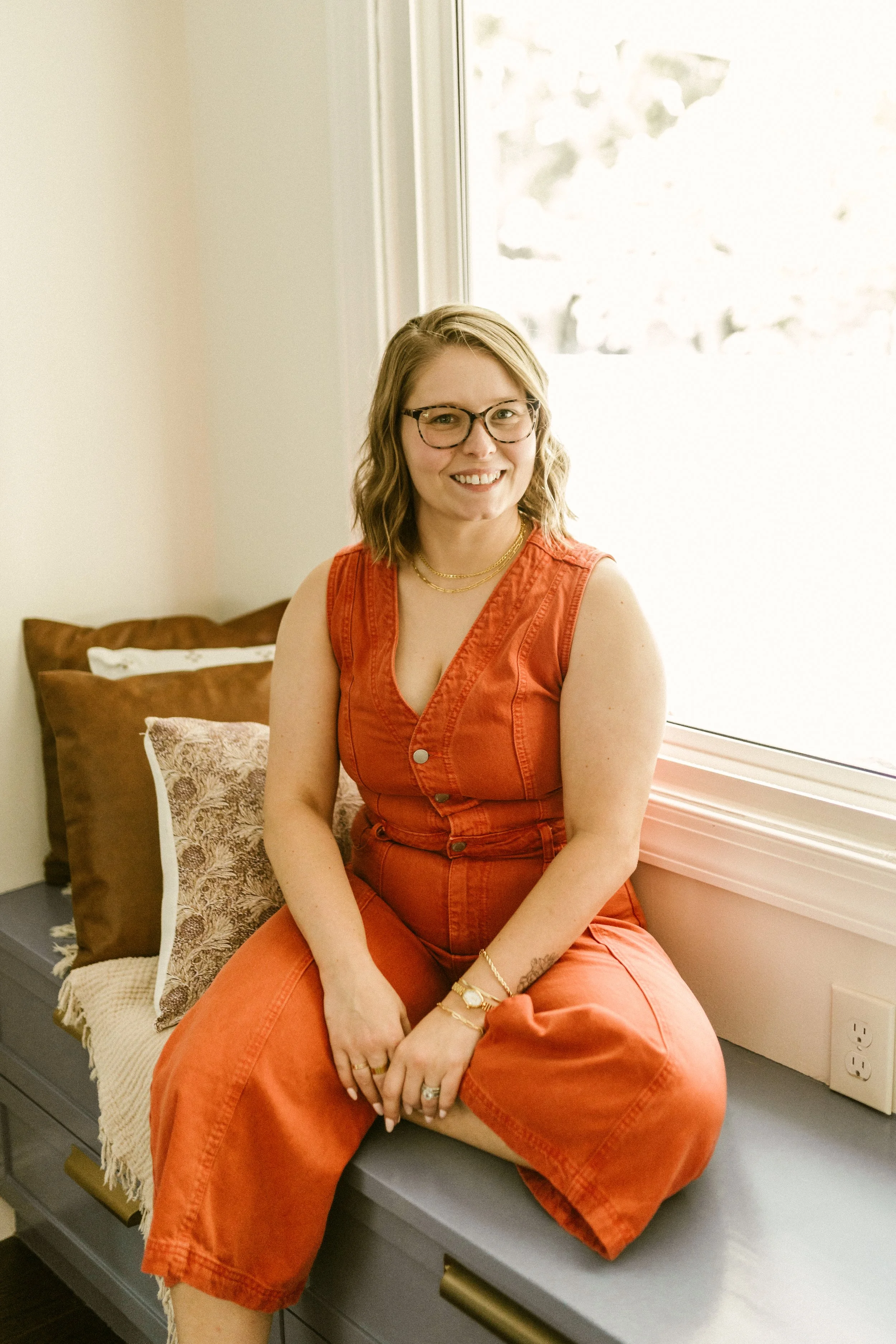 A woman with shoulder-length blonde hair, glasses, wearing an orange sleeveless jumpsuit, sitting on a windowsill with a beige wall and pillows behind her.