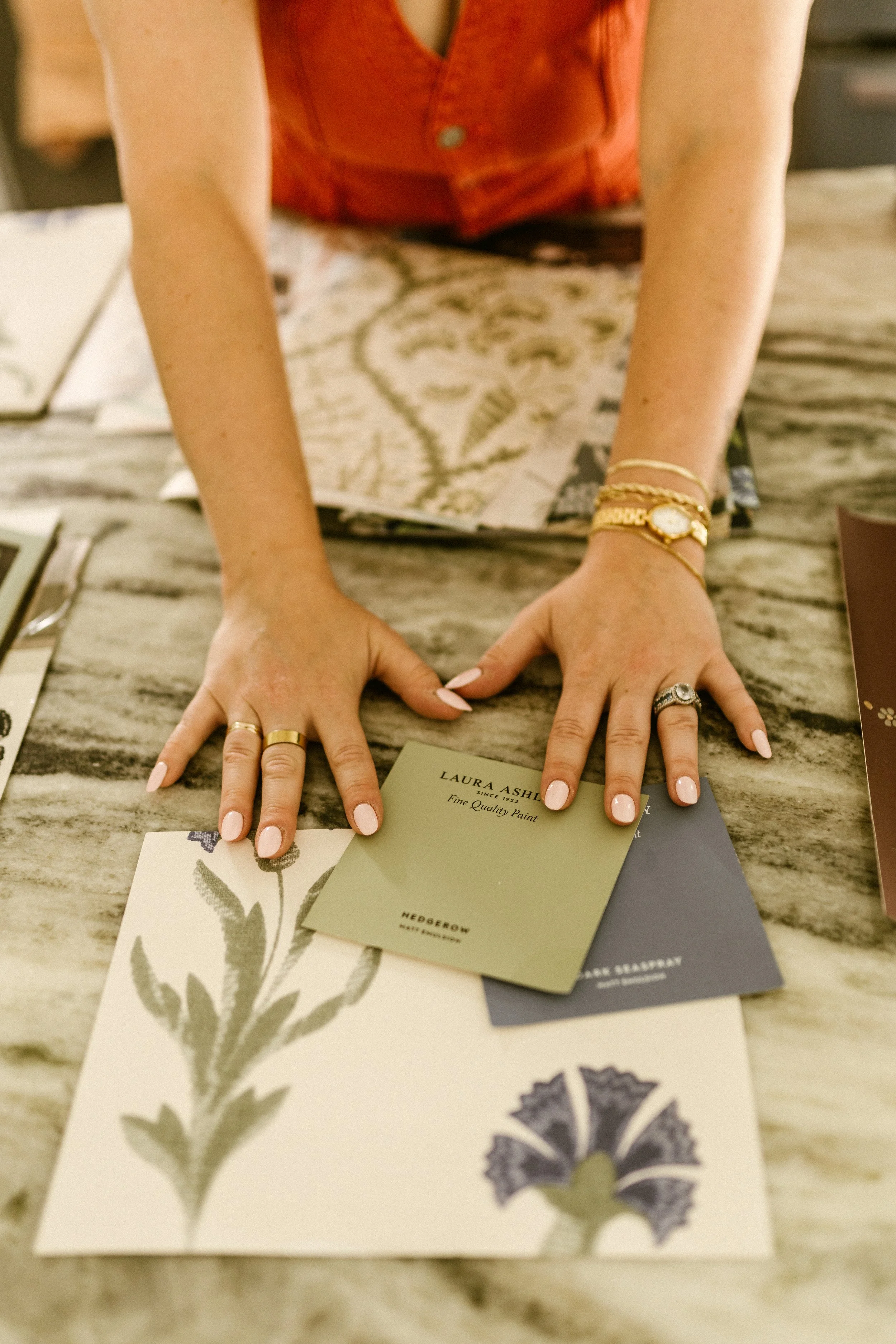 A woman resting her hands on a marble surface, displaying multiple rings and bracelets, with color sample cards and floral prints underneath.