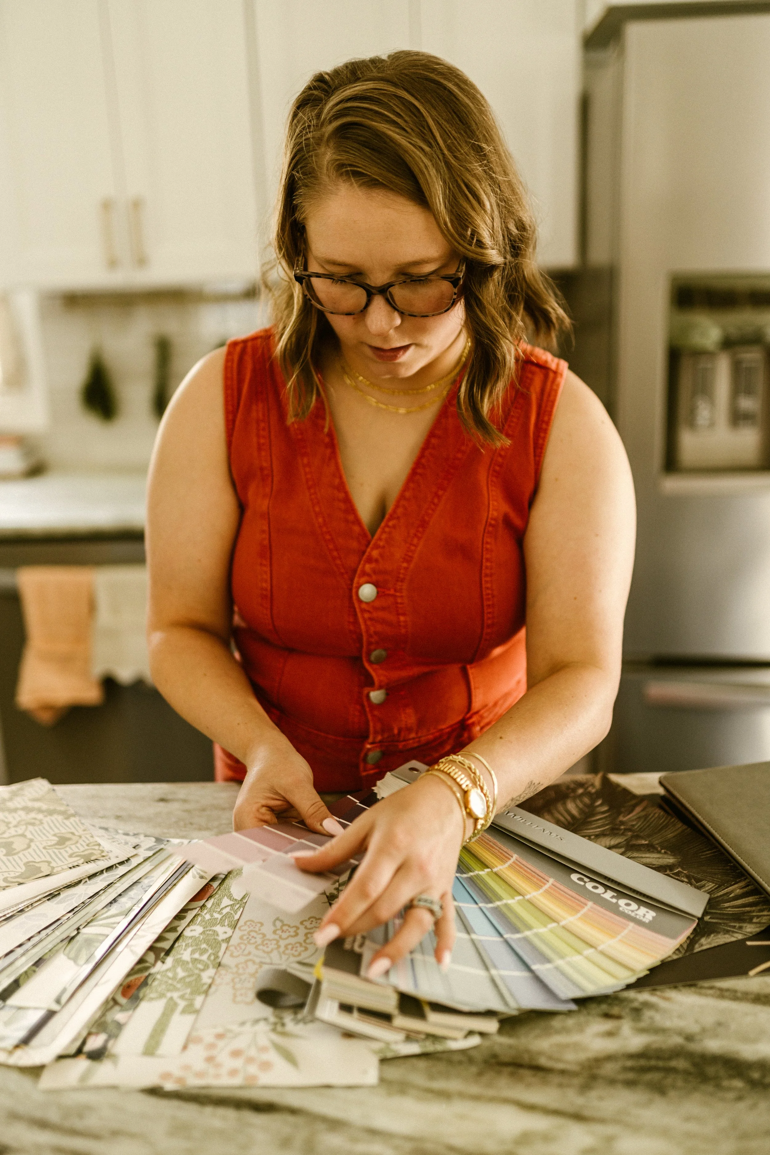 Woman with brown hair, glasses, wearing a sleeveless red top, looks down at a collection of paint color swatches on a wooden table, surrounded by decorative sample books.