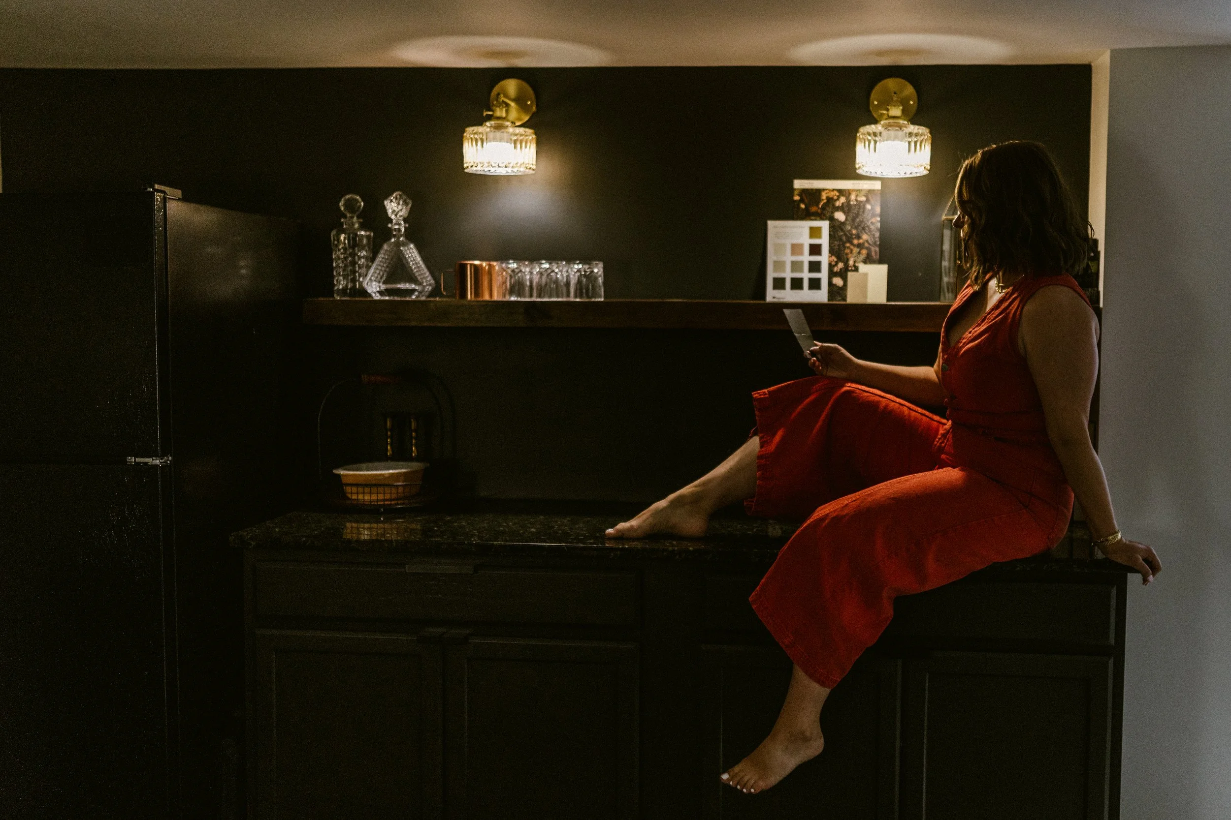 A woman in a red dress sitting on a kitchen countertop, looking at her phone, with one foot resting on the counter and the other dangling down.