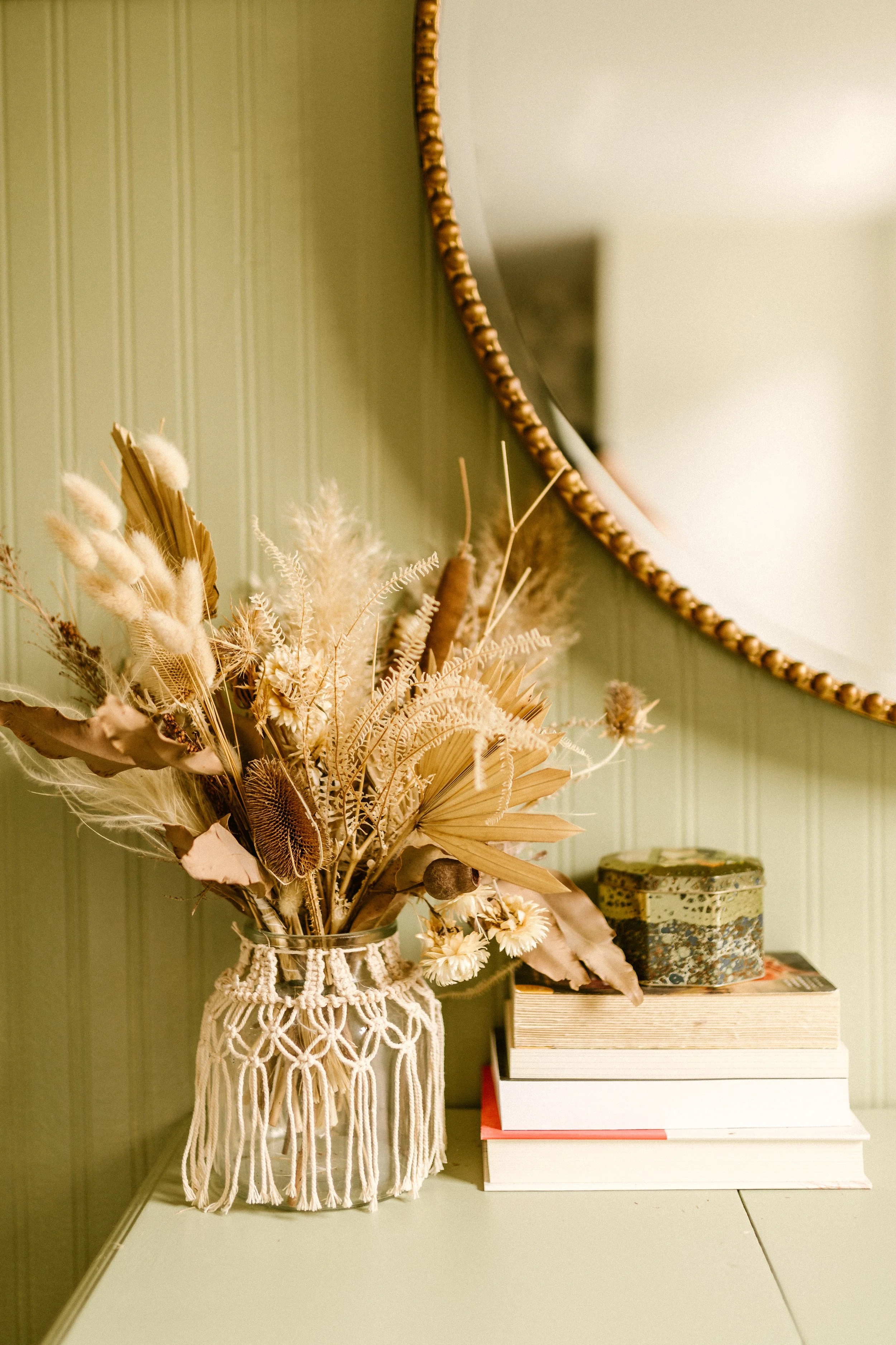 A decorative vase with dried flowers, placed on top of a stack of books and a vintage metal box in front of a mirror on a green wall.
