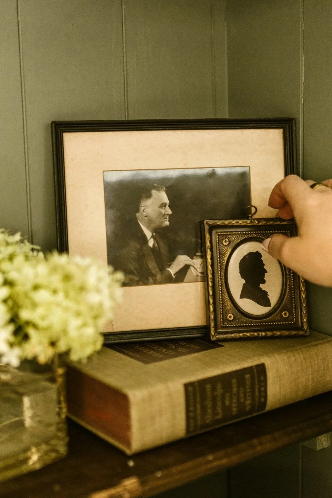 A close-up of a person's hand adjusting a small framed silhouette portrait on a shelf, with larger framed black-and-white photographs and antique books on a wooden surface.
