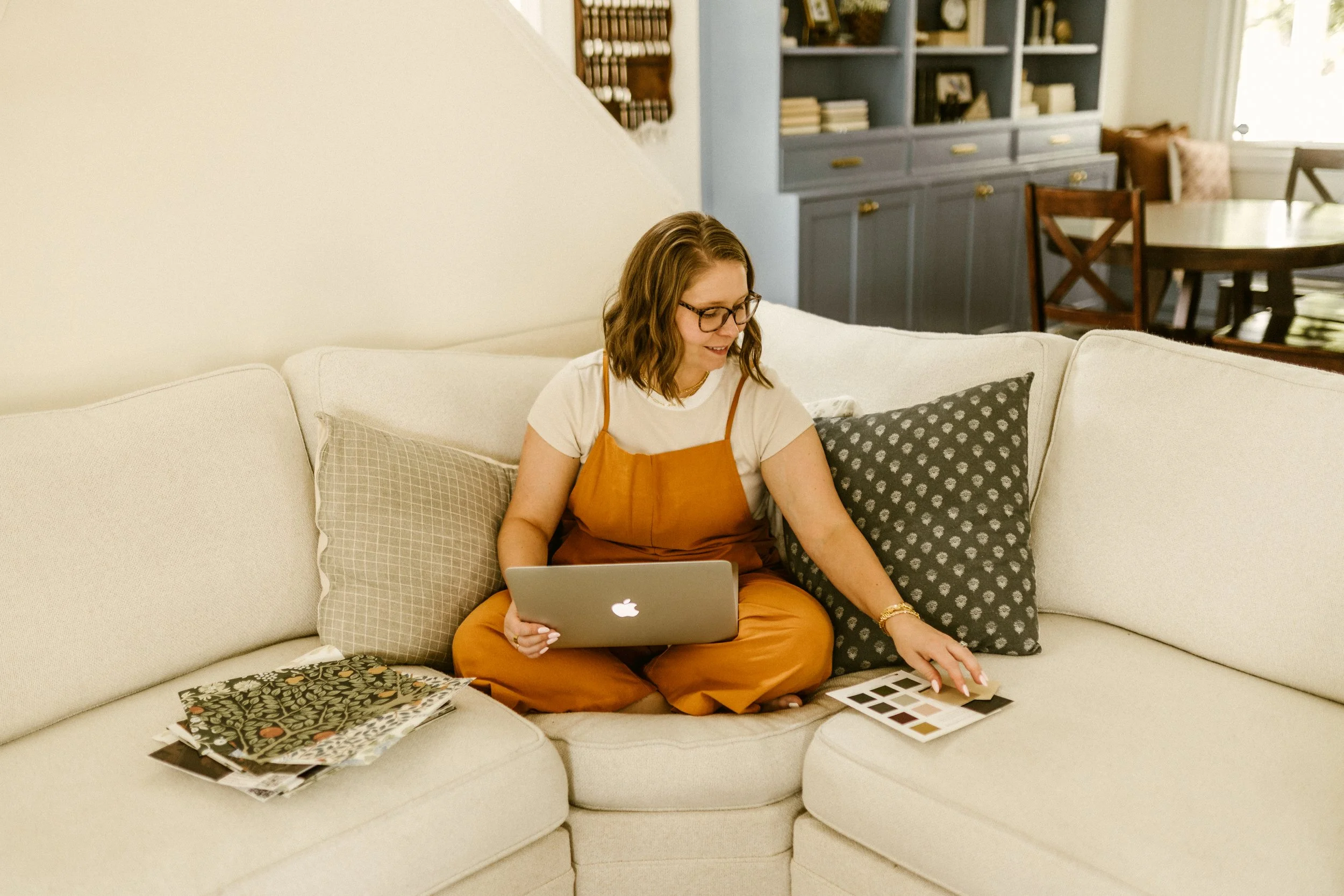 A woman with glasses and wavy hair sitting cross-legged on a white sofa, holding a silver MacBook and selecting color swatches from a sample book. There are color samples on the sofa and a dark green pillow behind her. In the background, there is a dark gray bookshelf and a dining area with a round table and wooden chairs.