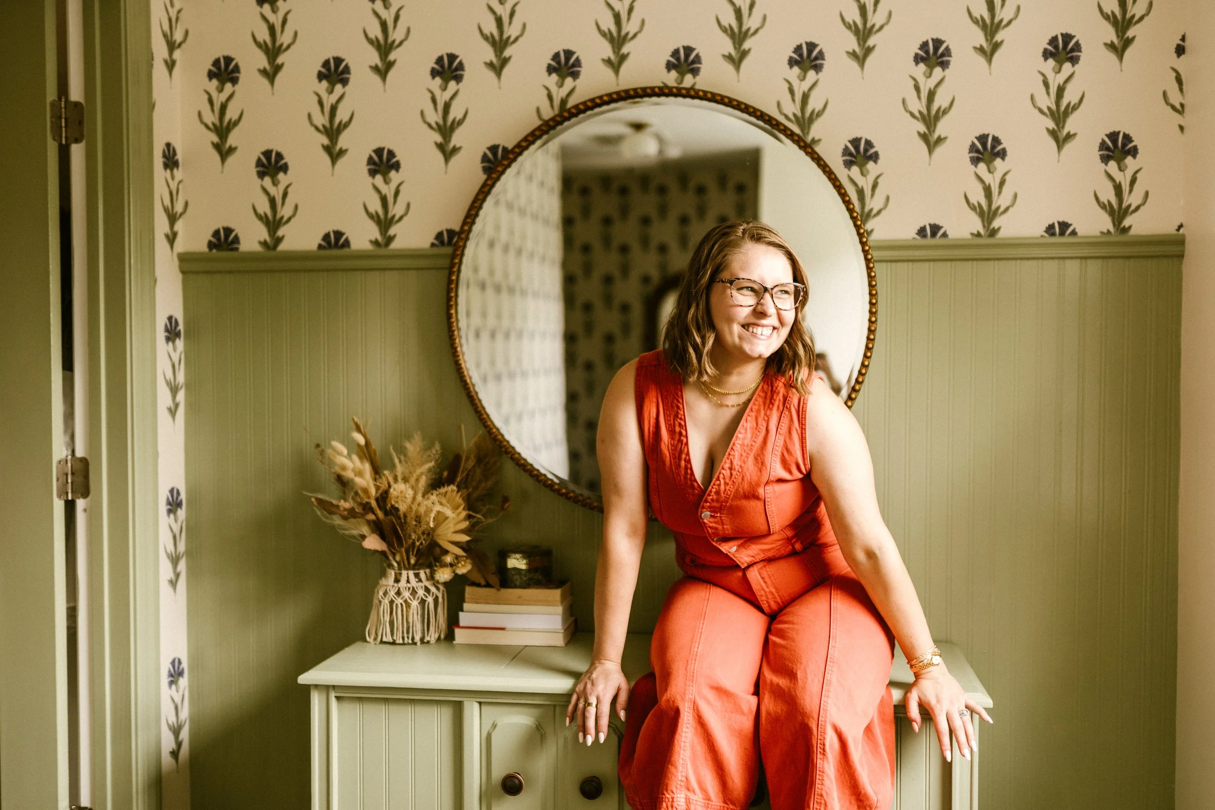 A woman with glasses and shoulder-length wavy hair in a red sleeveless jumpsuit, smiling and sitting on a green vintage dresser in a room with patterned wallpaper and a large round mirror behind her.