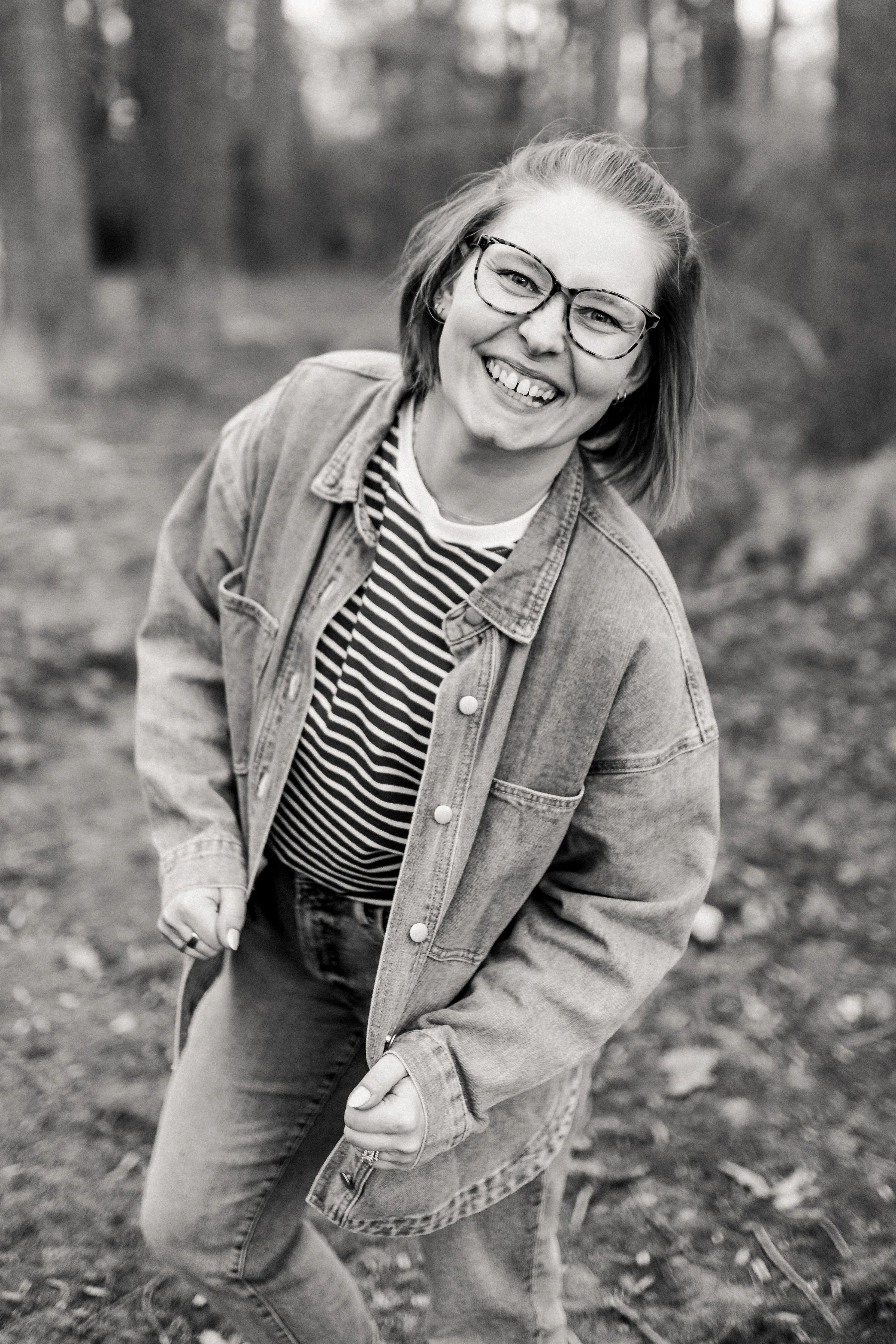 A woman smiling outdoors in a wooded area, wearing glasses, a striped shirt, a denim jacket, and jeans.