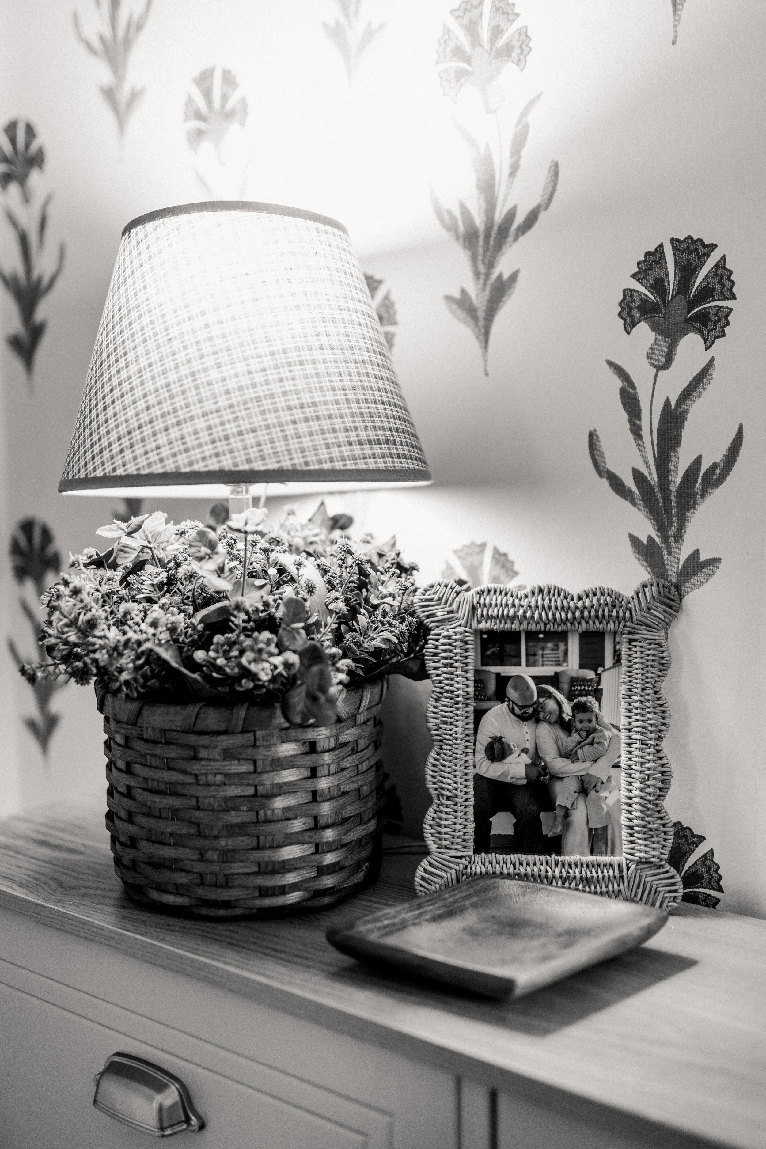 A houseplant with a lampshade on a wooden table, a picture frame with a photo, and a shallow dish, with floral wallpaper in the background.