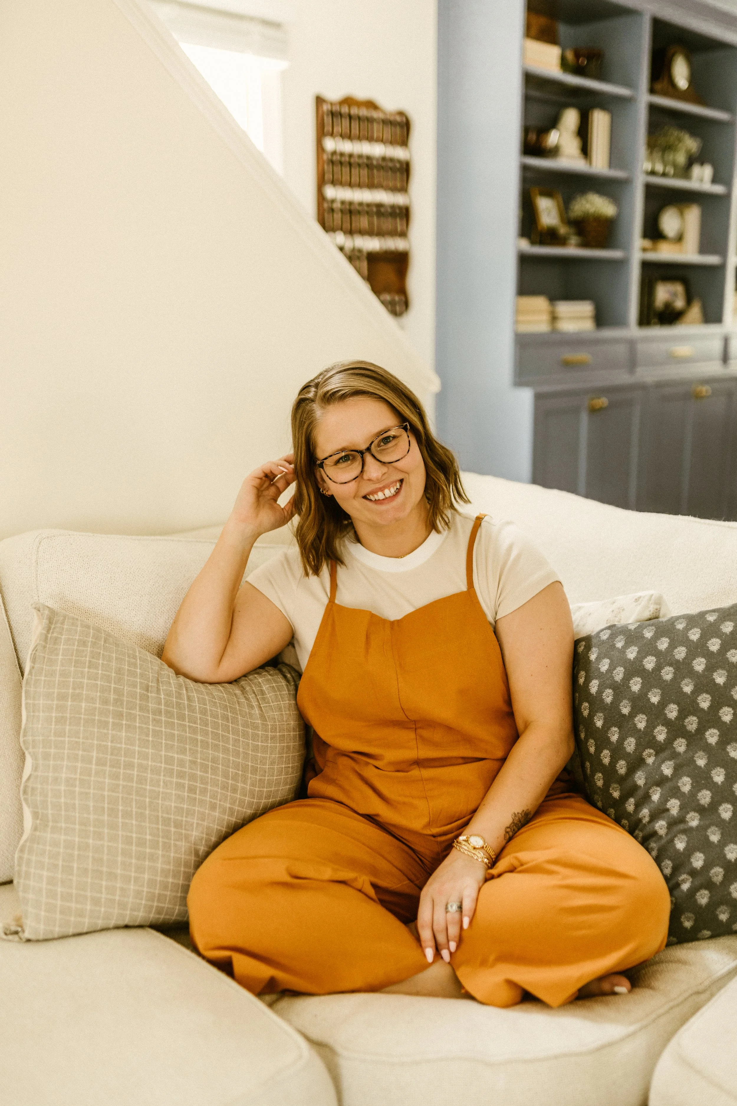 A woman with glasses, wearing a white t-shirt and orange overalls, sitting on a beige sofa, smiling, surrounded by cushions in a cozy living room.