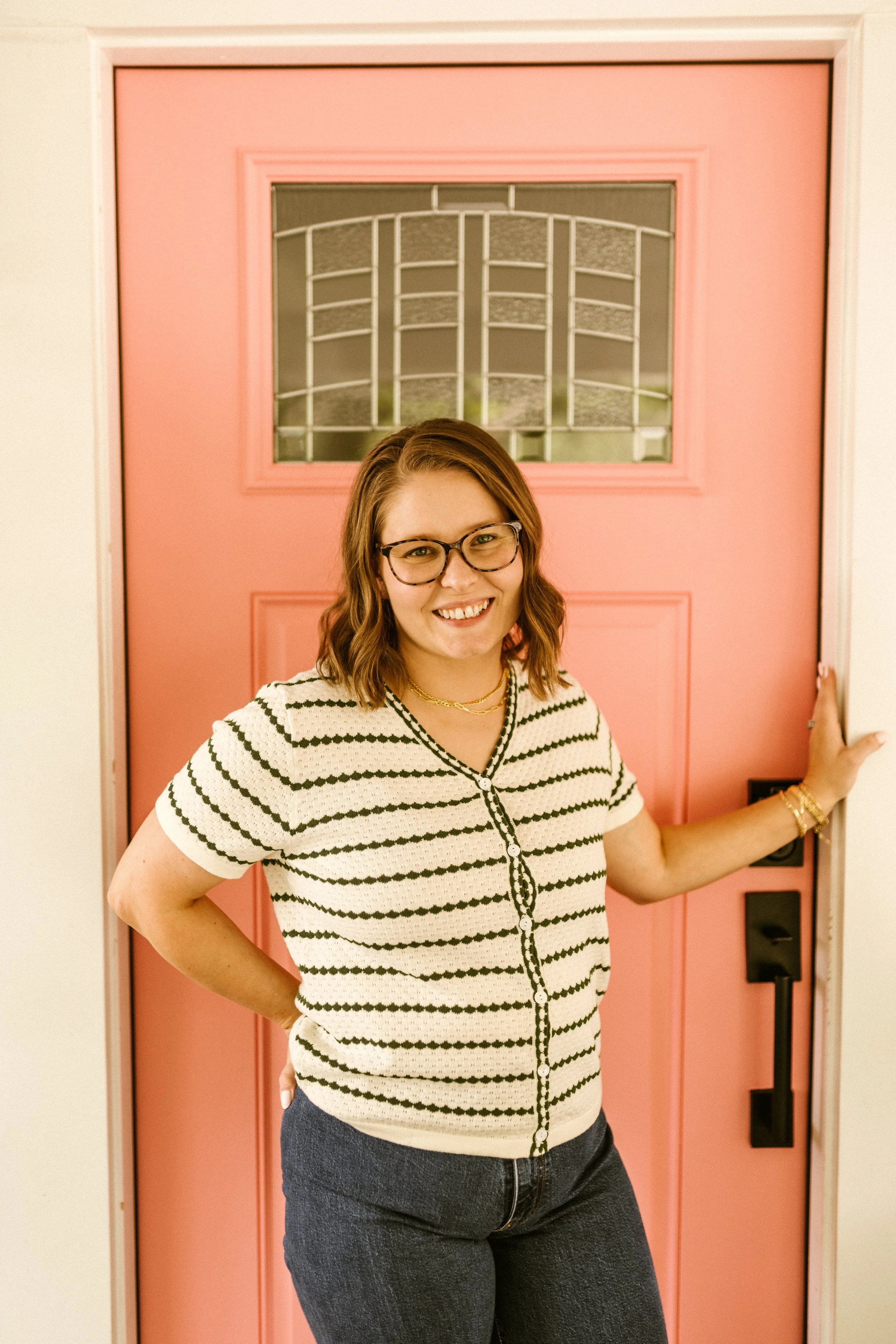 A woman with glasses and wavy brown hair smiling in front of a pink door, wearing a striped short sleeve top and dark jeans.