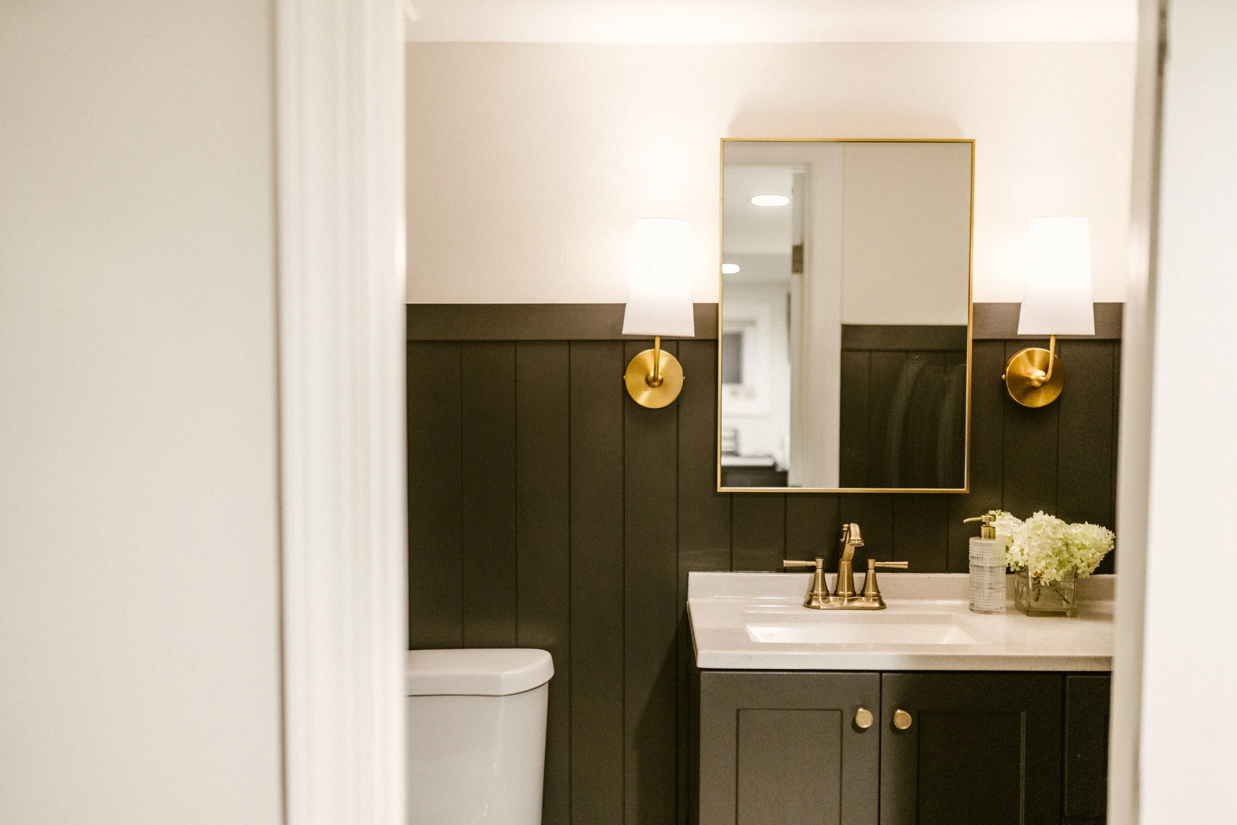 A bathroom viewed through a partially open door, featuring black paneled walls, a white toilet, a black vanity with a white countertop, a gold faucet, a rectangular mirror with a gold frame, wall-mounted lamps with white shades, and a vase of white flowers.